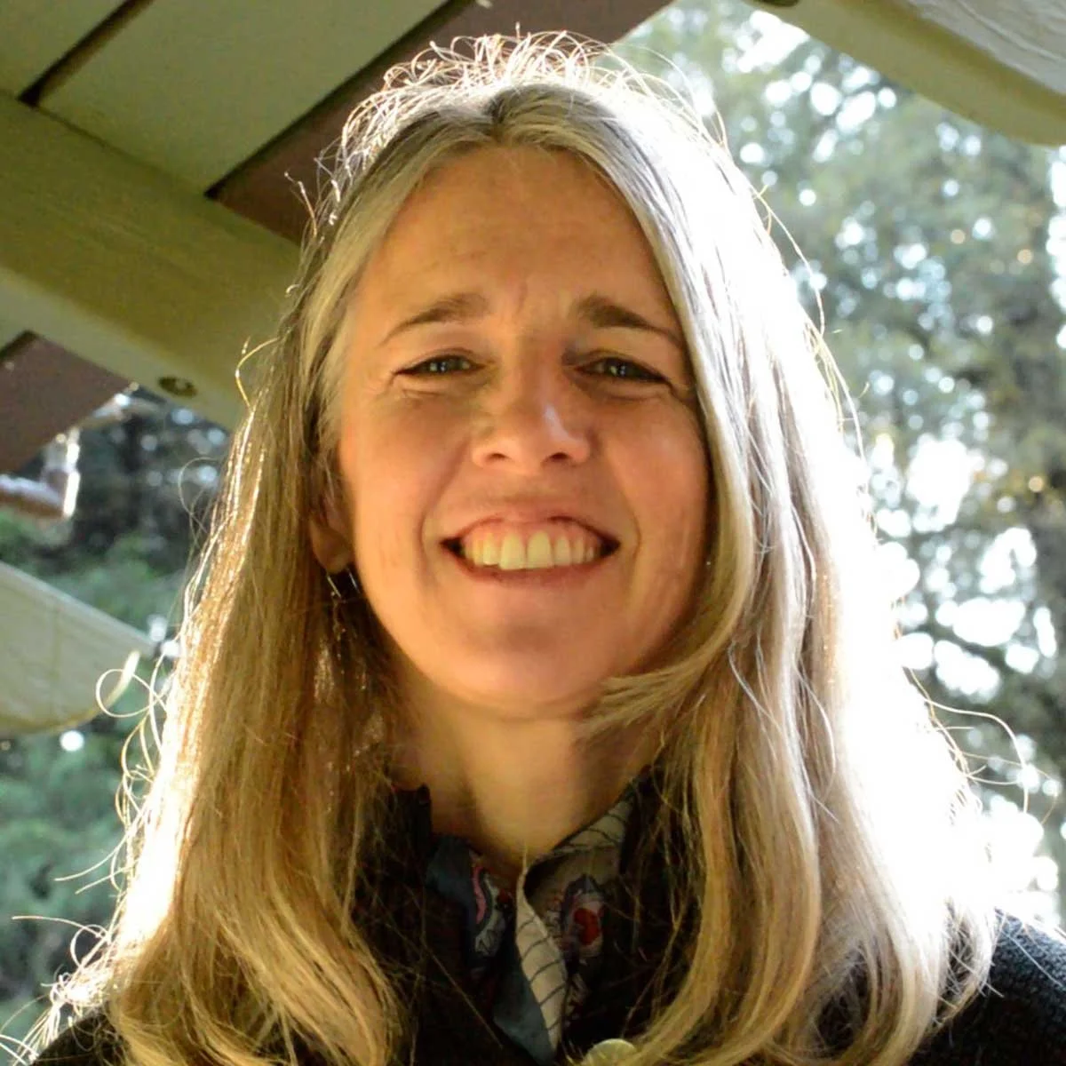 A smiling woman with long blonde hair outdoors in sunlight, with trees and part of a house visible in the background.