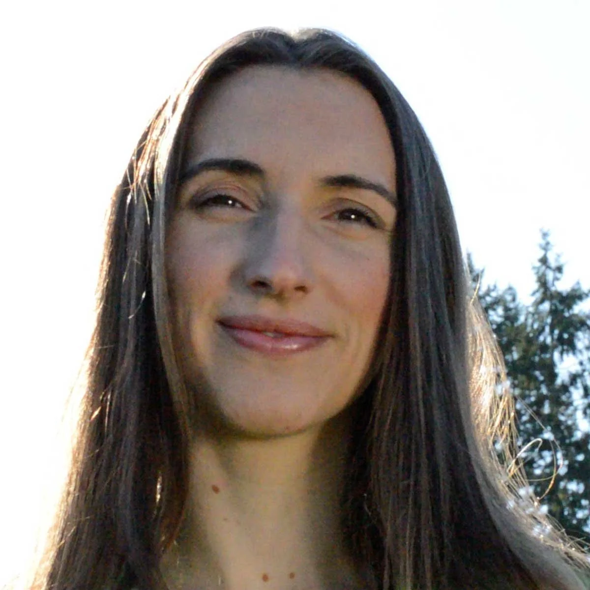 Close-up of a woman with long dark hair smiling outdoors with trees in the background and sunlight behind her.