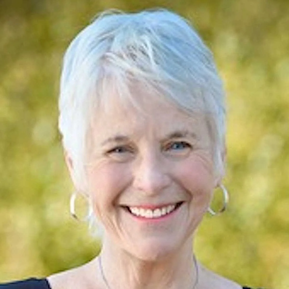 A middle-aged woman with short, light gray hair and blue eyes smiling outdoors, wearing hoop earrings and a dark top, with a blurred green background.