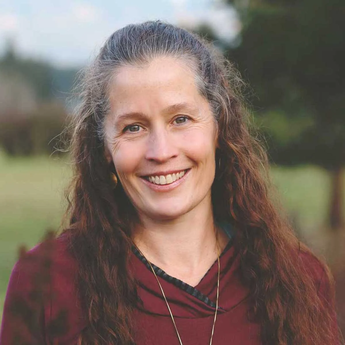 A smiling woman with long, curly brown hair, wearing a maroon top, outdoors with trees and grass in the background.