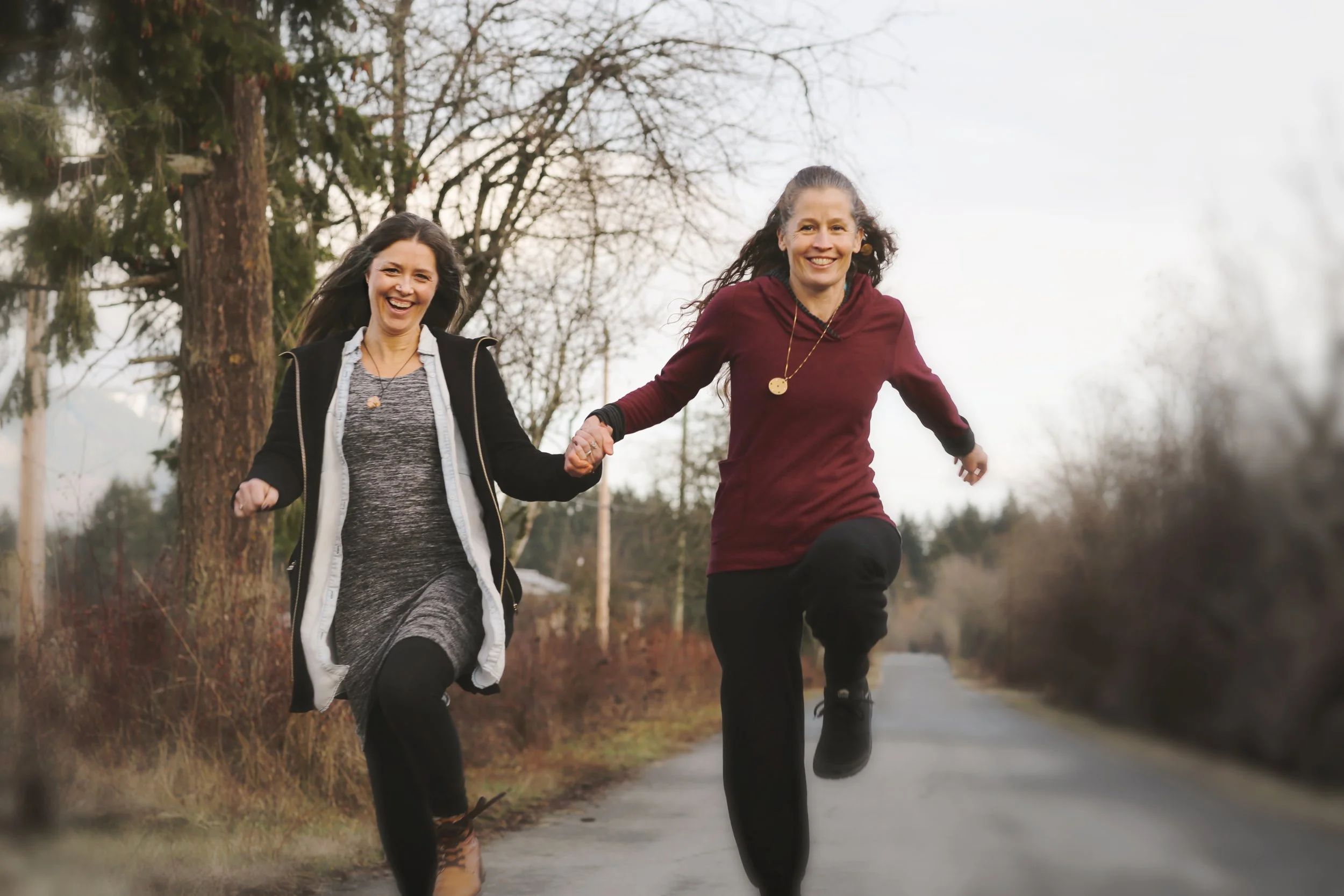 Two women running outdoors on a path, holding hands, with trees in the background on a cloudy day.