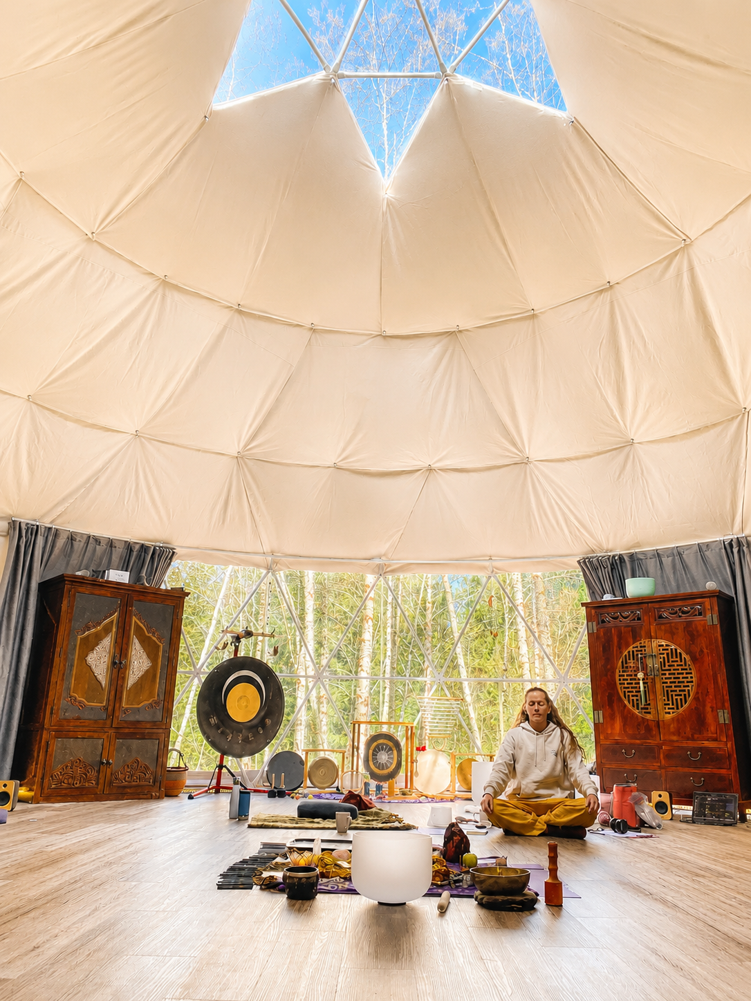Interior of a natural structure with a domed ceiling, large windows revealing trees outside, and a woman sitting cross-legged on the floor in a meditation pose surrounded by musical instruments and meditation supplies.