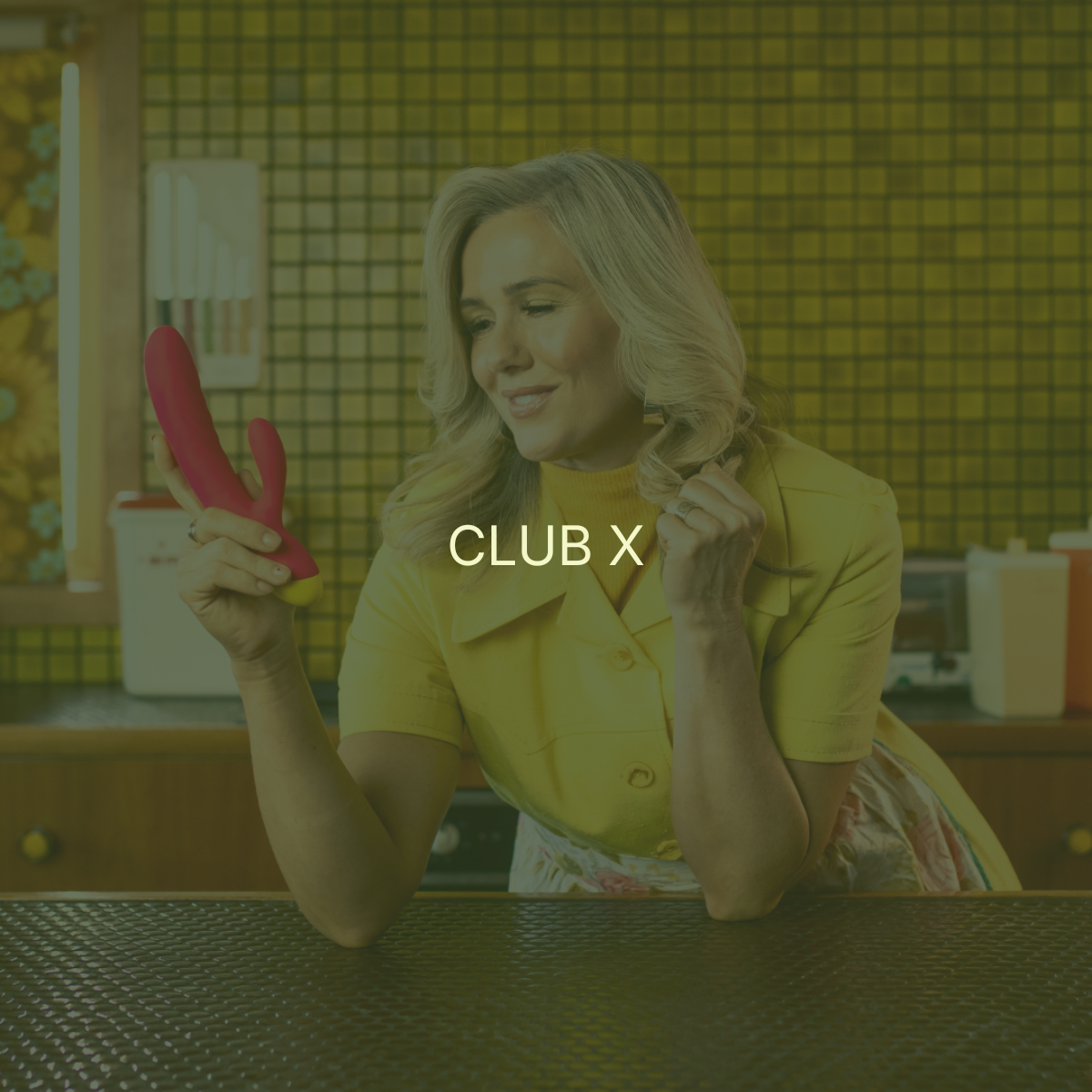A woman with blonde hair in a yellow shirt holding a pink and yellow vibrator, smiling in a colorful kitchen setting.