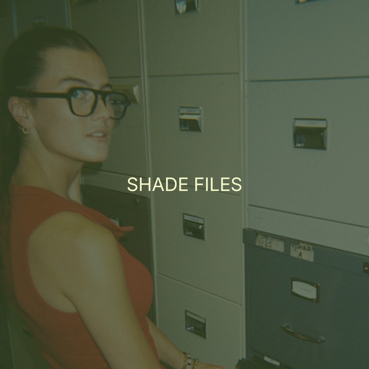 A woman with dark hair, glasses, and earrings standing next to grey mailboxes, with the text 'SHADE FILES' overlaid.