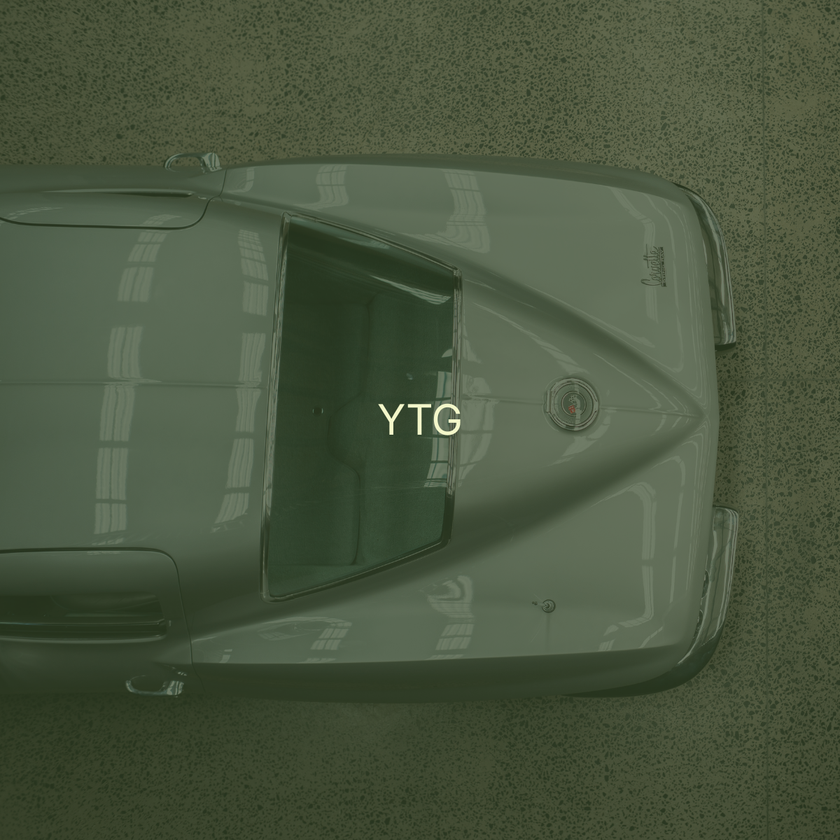 Top-down view of a vintage silver Corvette car parked in a garage, with the letters 'YTG' overlaid on the windshield.