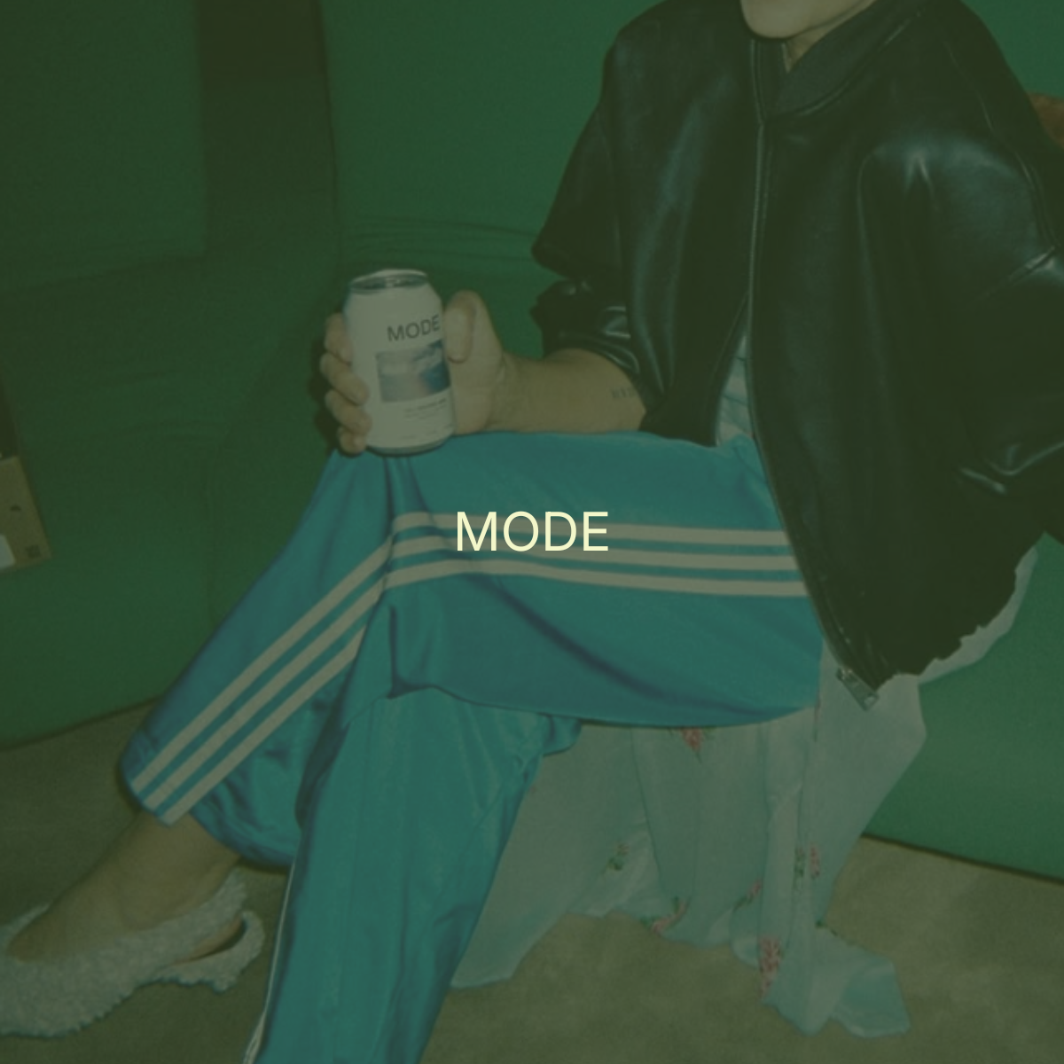 Person sitting on a box in a green room, wearing a black leather jacket, holding a can of MODE beverage, with the word 'MODE' overlayed on the image.