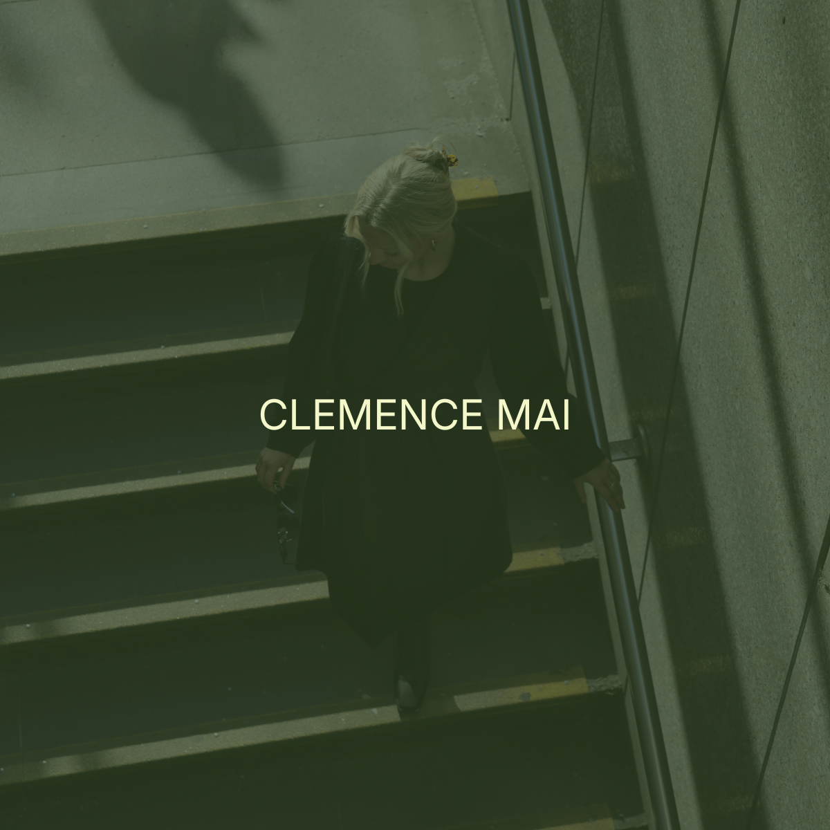 A woman with blonde hair walking down a staircase, holding a phone in her right hand and a purse in her left, wearing a black coat, in an indoor setting with concrete walls and stairs. The text 'Clemence Mai' overlays the image.