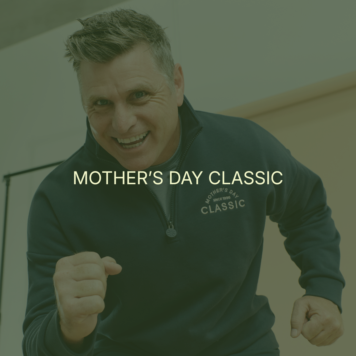 A smiling man with short hair, wearing a dark zip-up jacket with the words 'Mother's Day Classic' on it, appears excited or energetic. He is indoors, possibly at a celebratory or athletic event.