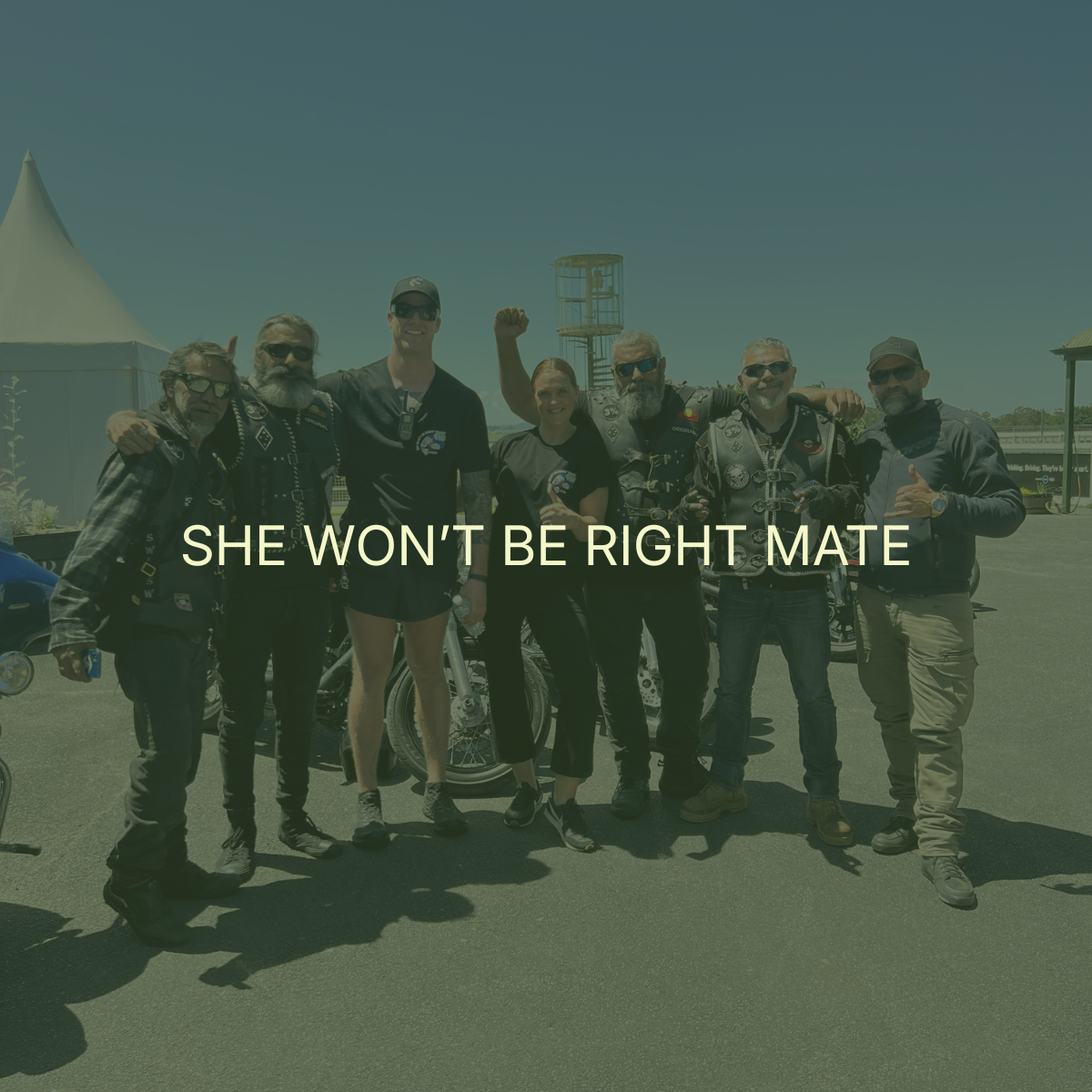 Group of six people standing outdoors, some wearing motorcycle vests, with motorcycles and a tower in the background, under a clear sky.