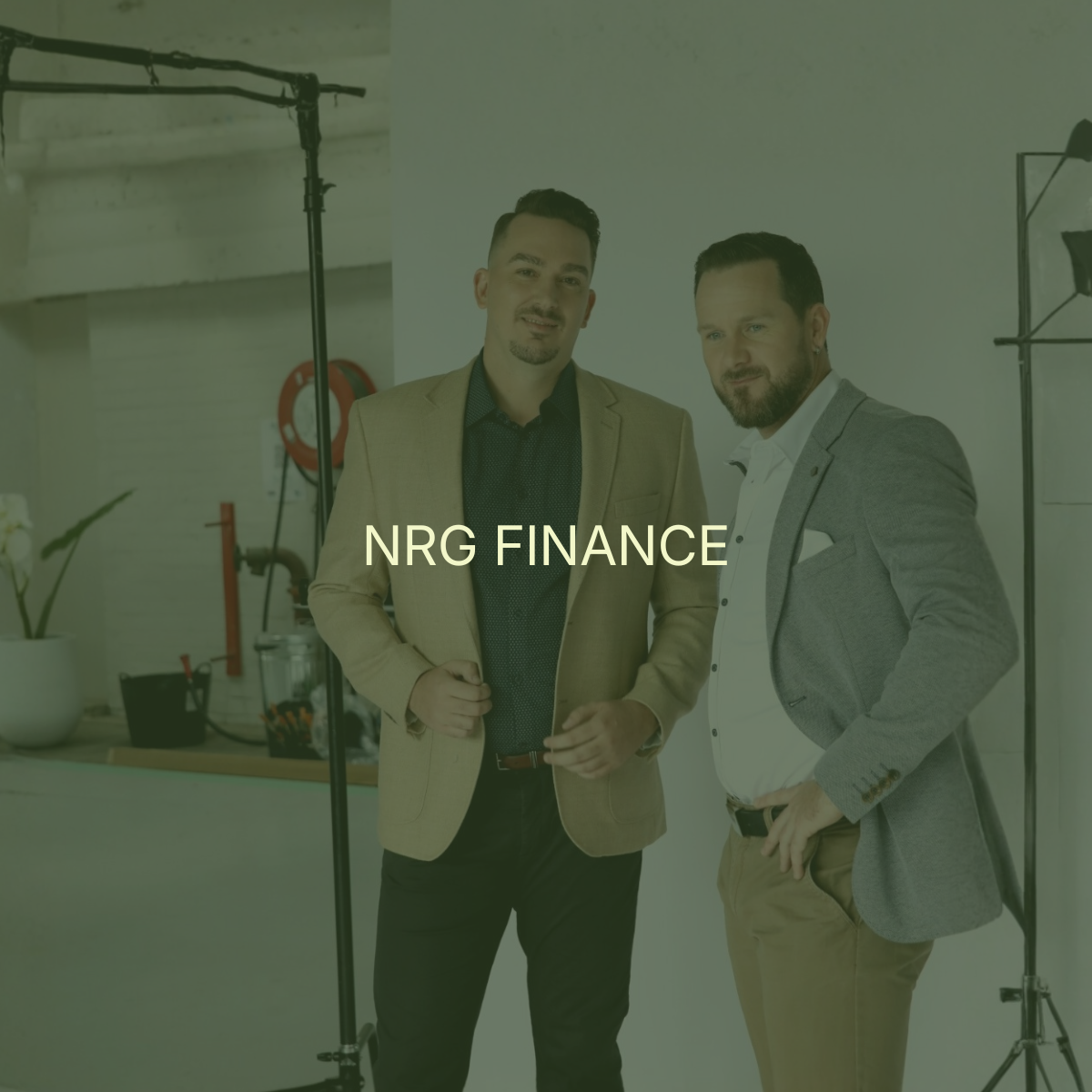 Two men in business attire standing together in a modern office space with lighting equipment visible in the background, under the text 'NRG FINANCE'.