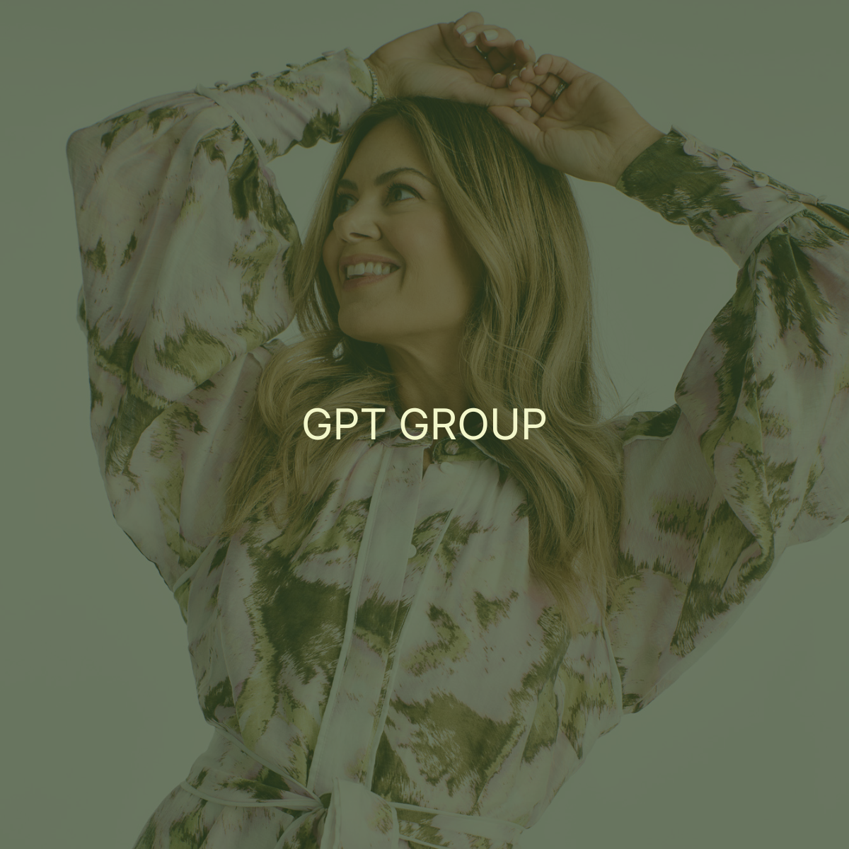 A woman smiling with her hands on her head, wearing a long-sleeved, floral or abstract patterned blouse.