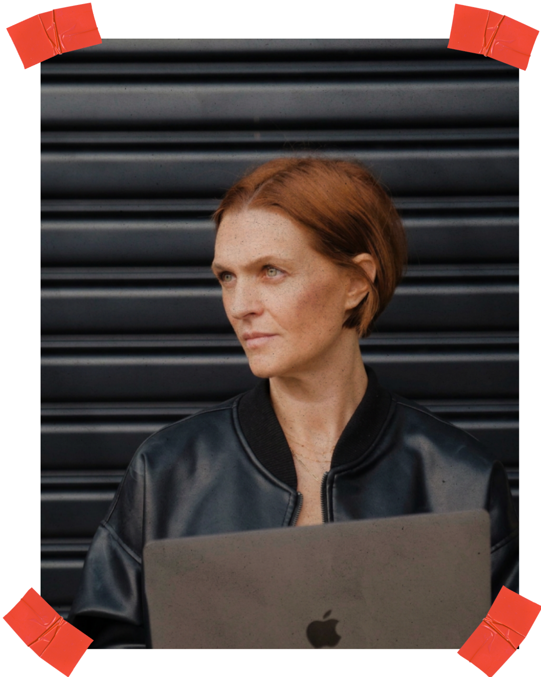 A woman with short red hair, wearing a black leather jacket, looking to the side while holding a silver MacBook laptop, standing in front of a black metal roller shutter.