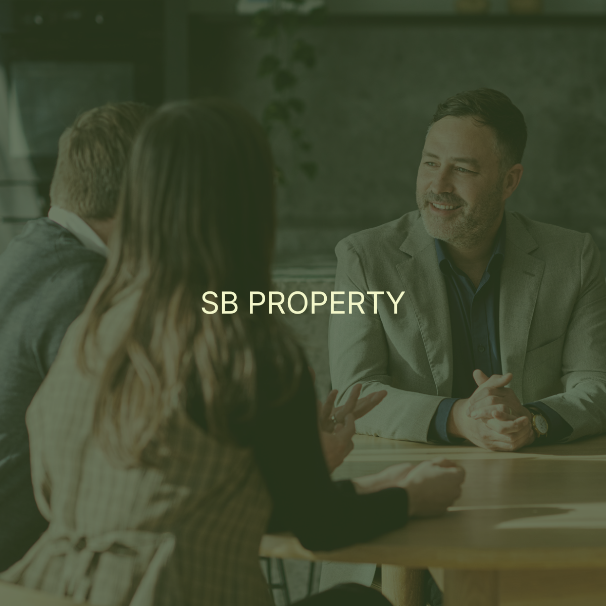 A business meeting with three people sitting around a table, a man in a gray suit talking to a woman with long hair and glasses. The text overlay reads 'SB PROPERTY'.