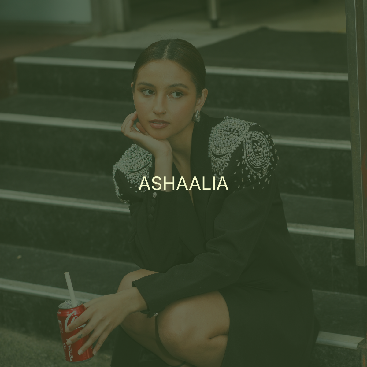A young woman with dark hair styled back, sitting on steps, wearing a black outfit with intricate, embellished shoulders, holding a red soda can with a straw, and gazing thoughtfully to the side. The word "ASHAALIA" is overlaid in the center.