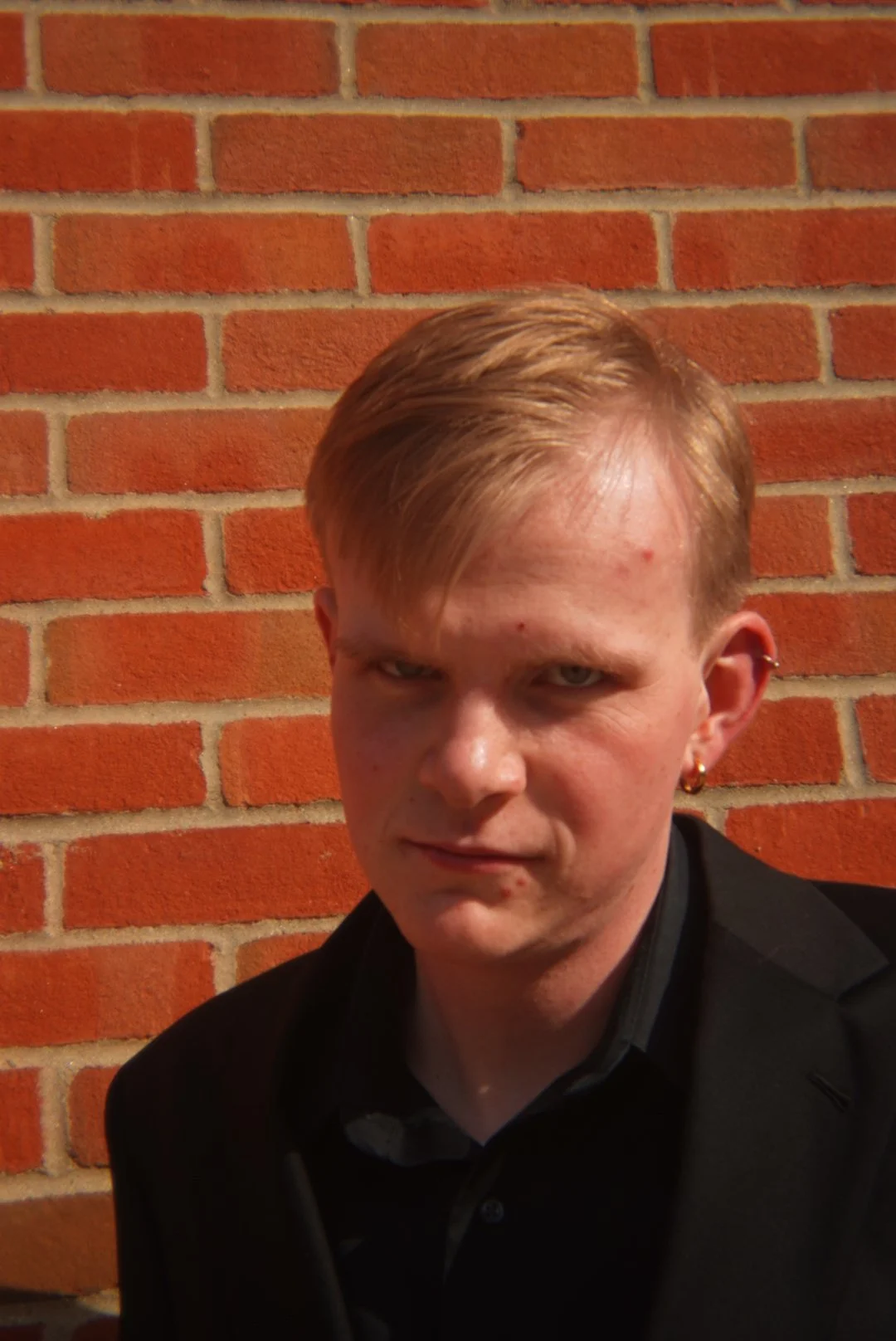 A young man with red hair, dressed in a black coat and shirt, standing against a red brick wall.