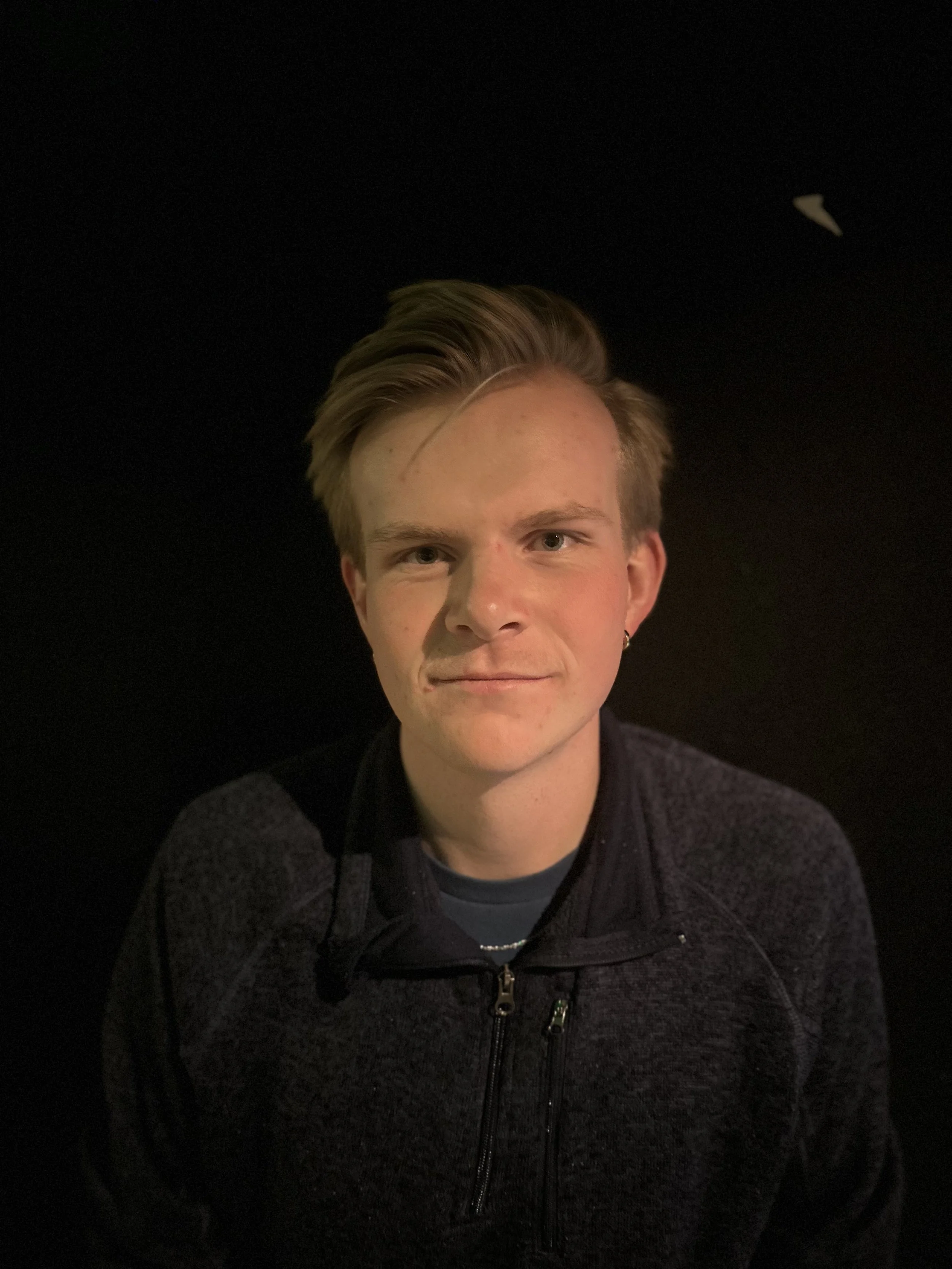 Young man with short blond hair smiling and wearing a black zip-up jacket, in a dark background.