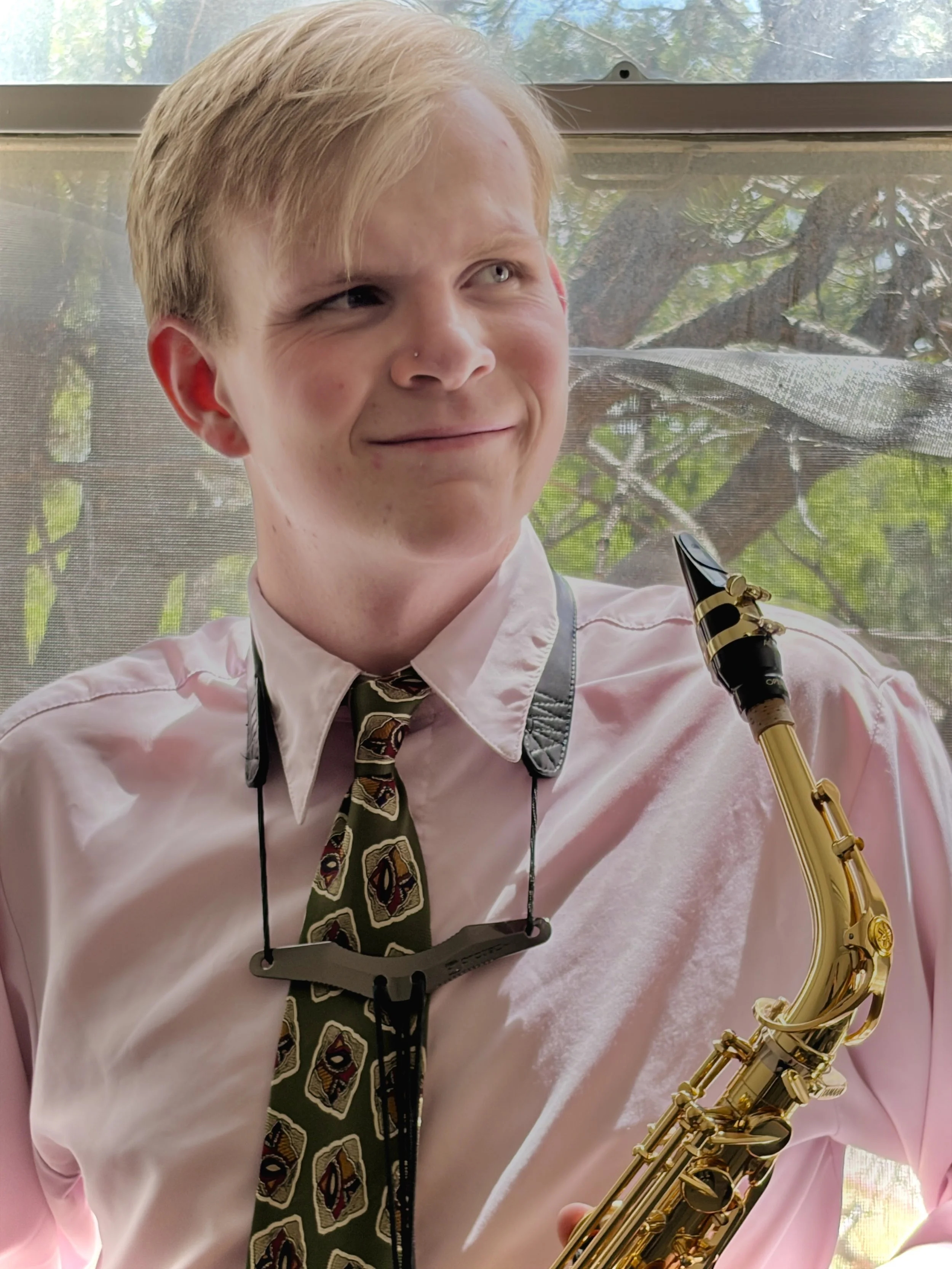 A young person with blonde hair and a piercing on the nose, wearing a white shirt and a patterned tie, is holding a gold-colored saxophone. They are standing in front of a window with a view of green trees outside.