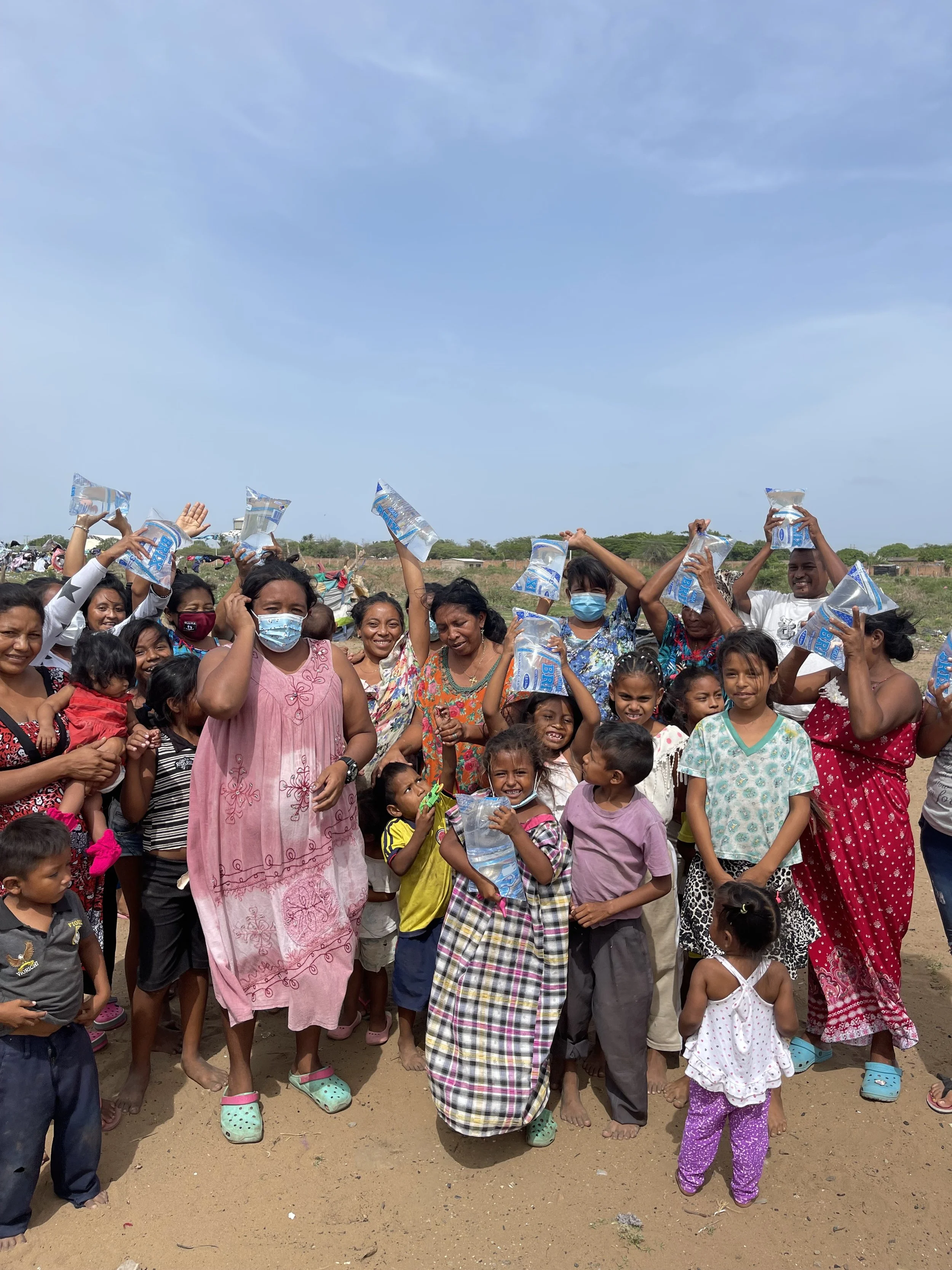 A large group of smiling people, including children and adults, gathered outdoors on a dirt ground under a blue sky, some holding plastic water bags, with some wearing face masks and colorful clothing.