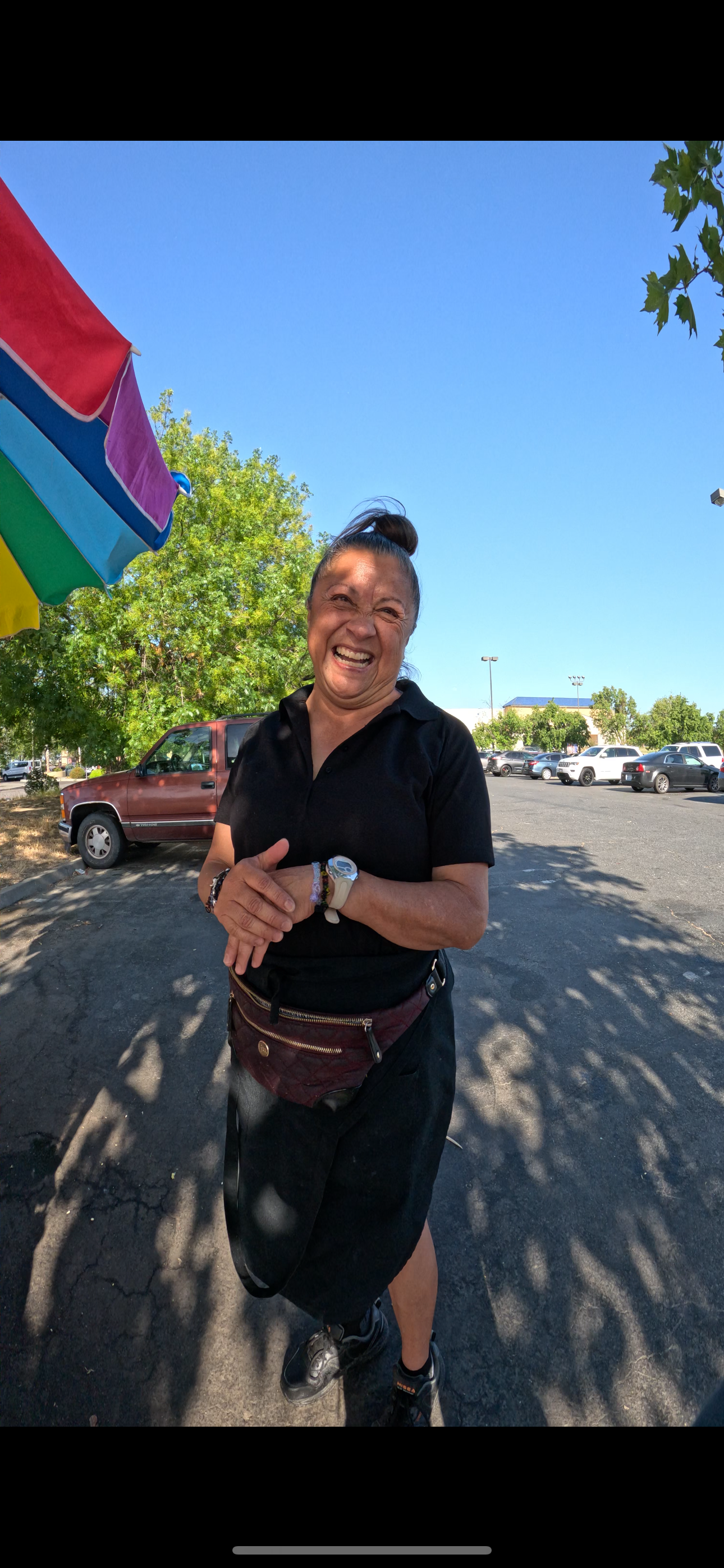 A woman smiling and laughing outdoors under a bright blue sky, standing in a parking lot with cars in the background, wearing a black polo shirt, a fanny pack, and sports shoes.