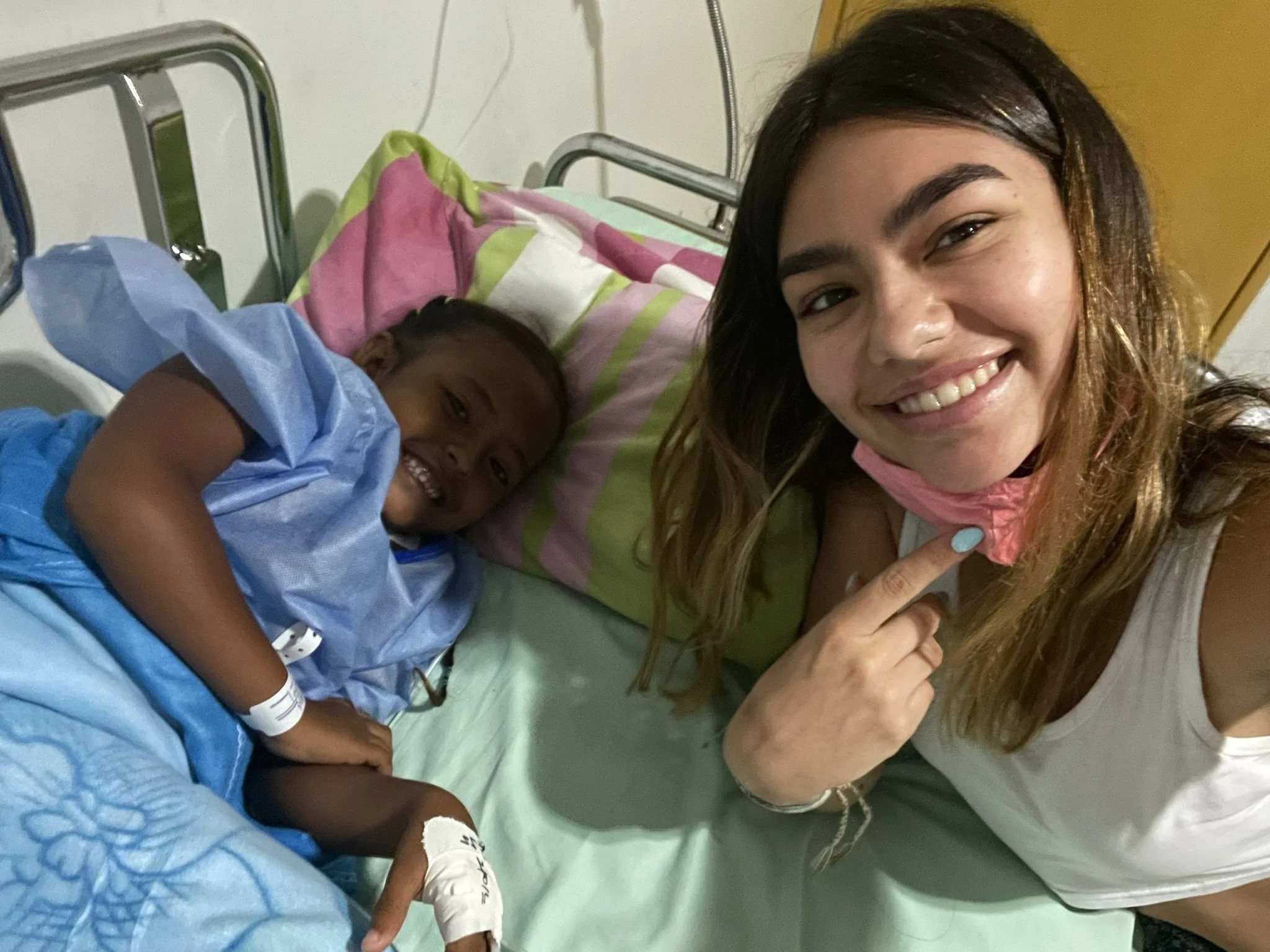 A woman with long hair smiling and pointing at a young boy lying on a hospital bed, who is wearing a hospital gown and has an IV in his arm, in a hospital room.
