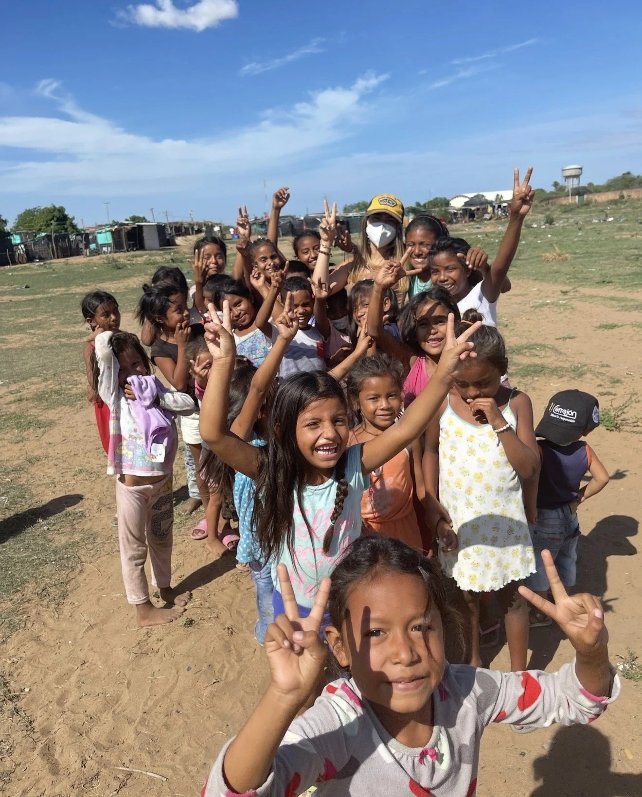 A group of children and a woman wearing a mask, standing outdoors in a rural area under a blue sky. The children are smiling and making peace signs.
