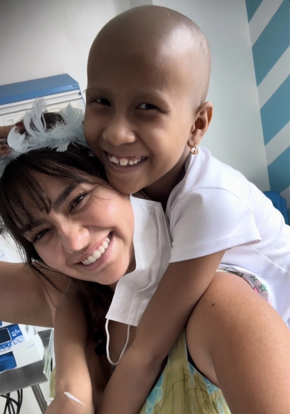 A woman with brown hair and light skin, wearing a white top, is smiling and taking a selfie with a young girl who has a shaved head, light skin, earrings, and is smiling. The girl is wearing a white shirt and earrings. They appear happy and are in a medical setting with medical equipment in the background.