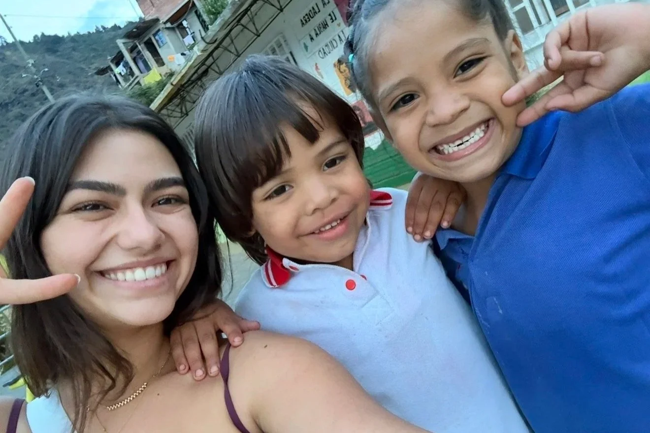 A young woman and two boys smiling and making peace signs with their hands outdoors.