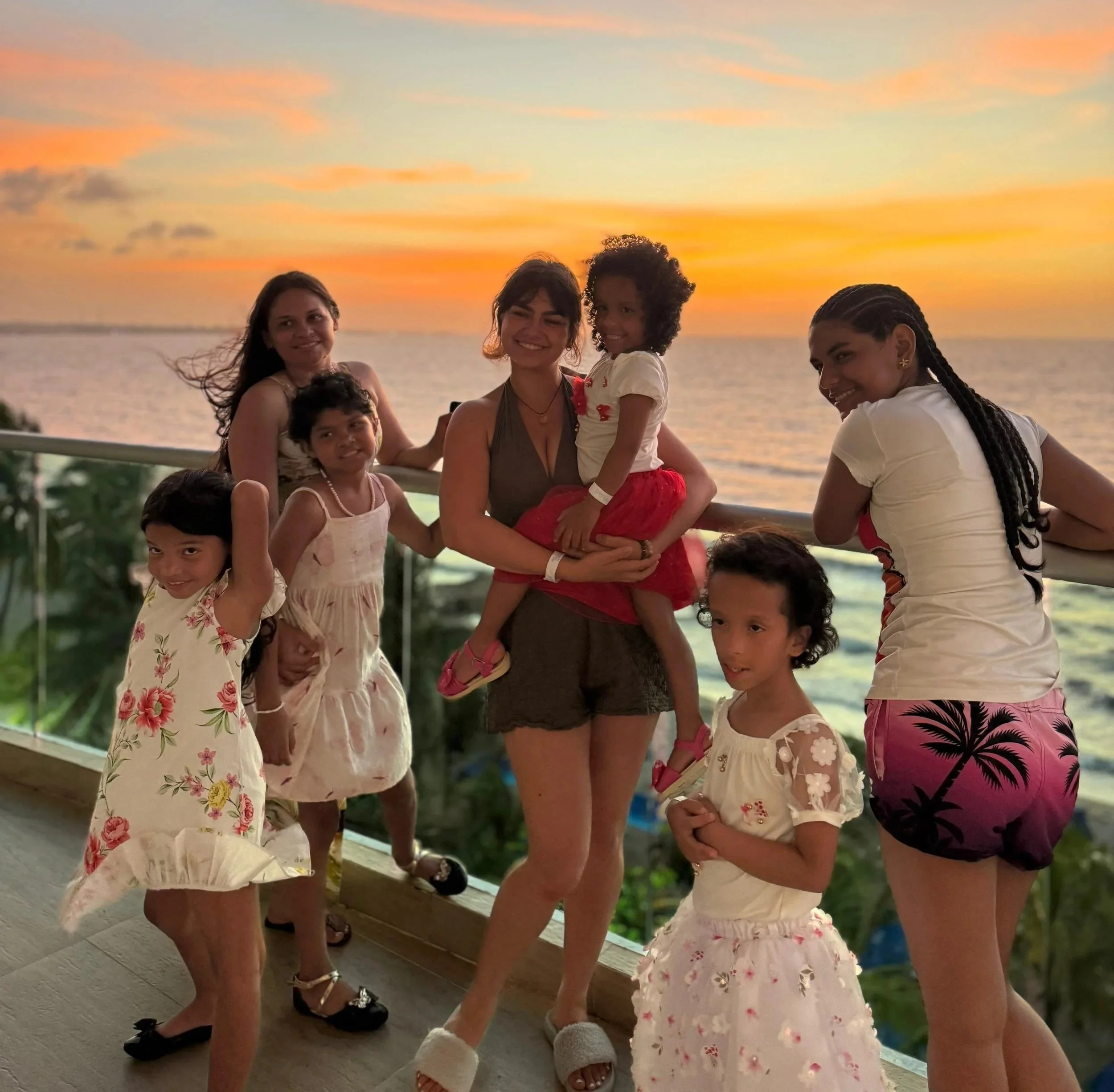 A group of six young girls standing and sitting on a deck at sunset, dressed in summer clothes, with water and a colorful sky in the background.
