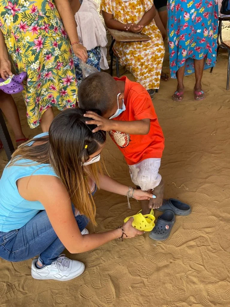 A woman crouching on sandy ground helping a boy put on Crocs shoes, with other women standing around and watching. The boy and woman are wearing face masks, and the boy is wearing a red shirt. The women are wearing colorful, floral maxi dresses.