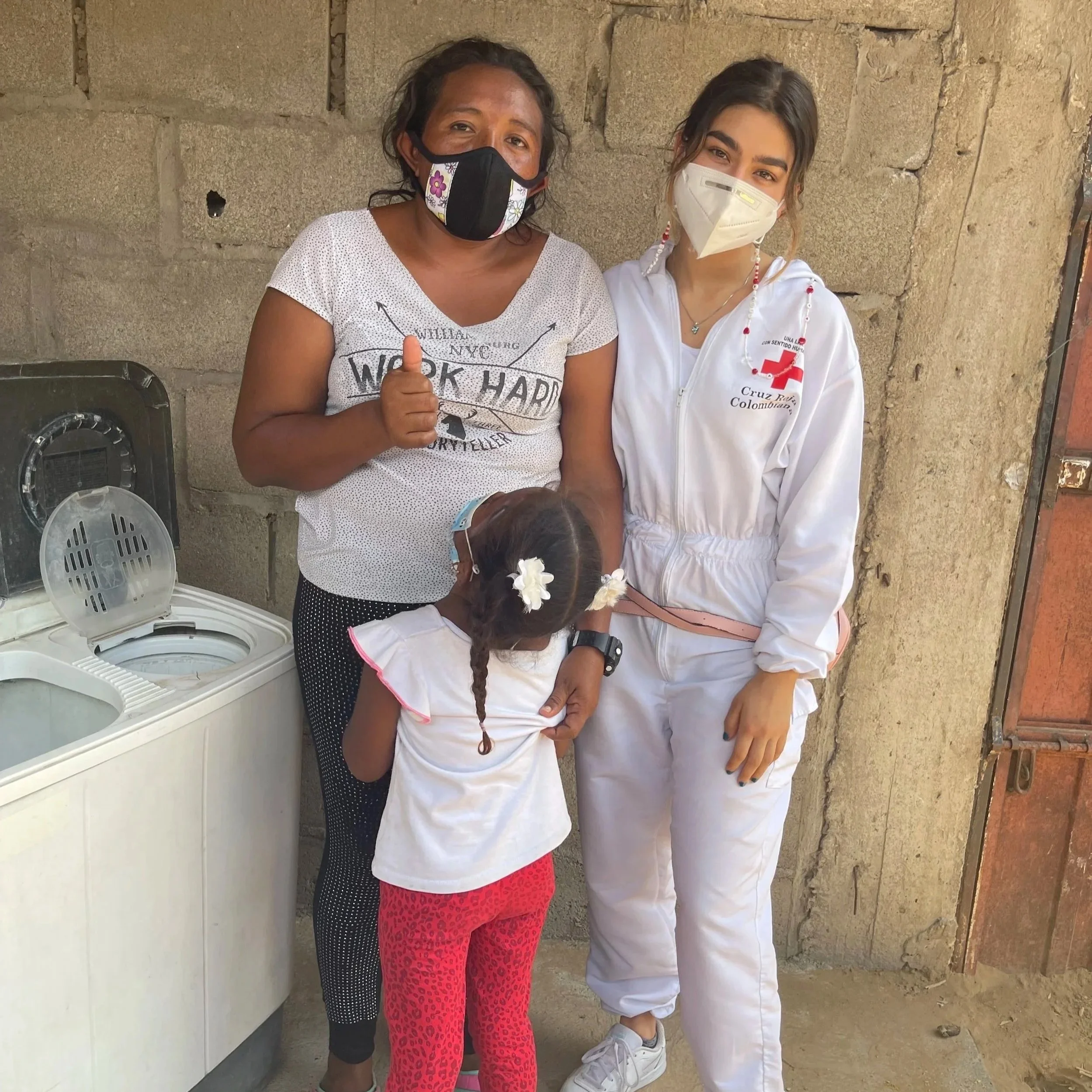 Two women wearing face masks, standing side by side against a brick wall. One woman is in casual clothes giving a thumbs-up, and the other is in a white uniform with aRed Cross patch.