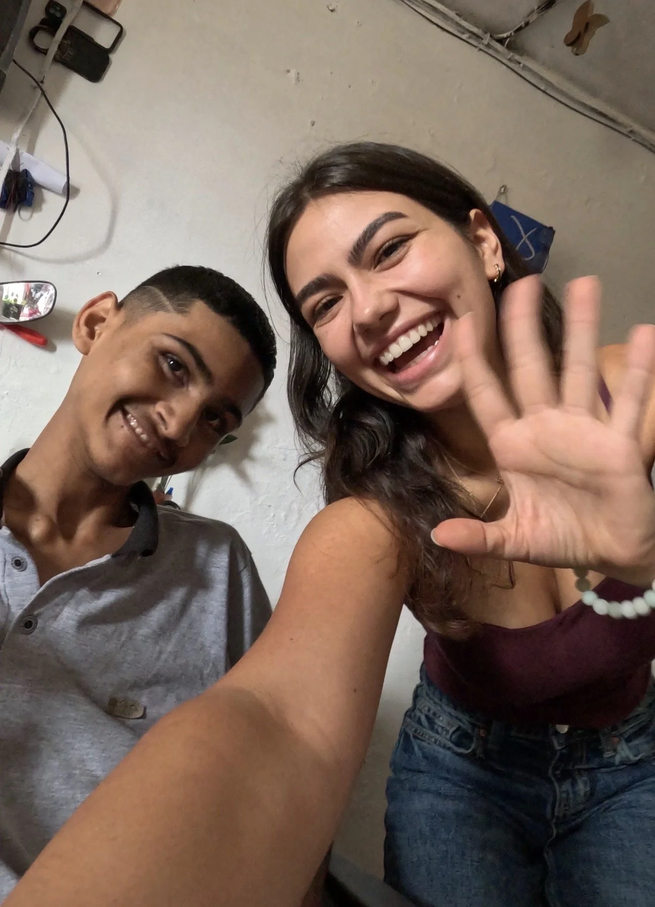 Two young women taking a selfie in an indoor setting, smiling and waving at the camera.