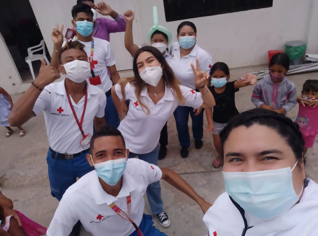 Group of medical volunteers and children wearing masks, some with Red Cross shirts, taking a selfie outdoors.