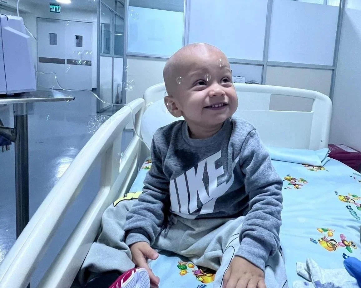 A young child with a shaved head sitting up in a hospital bed, smiling and looking to the side, with a hospital room background.