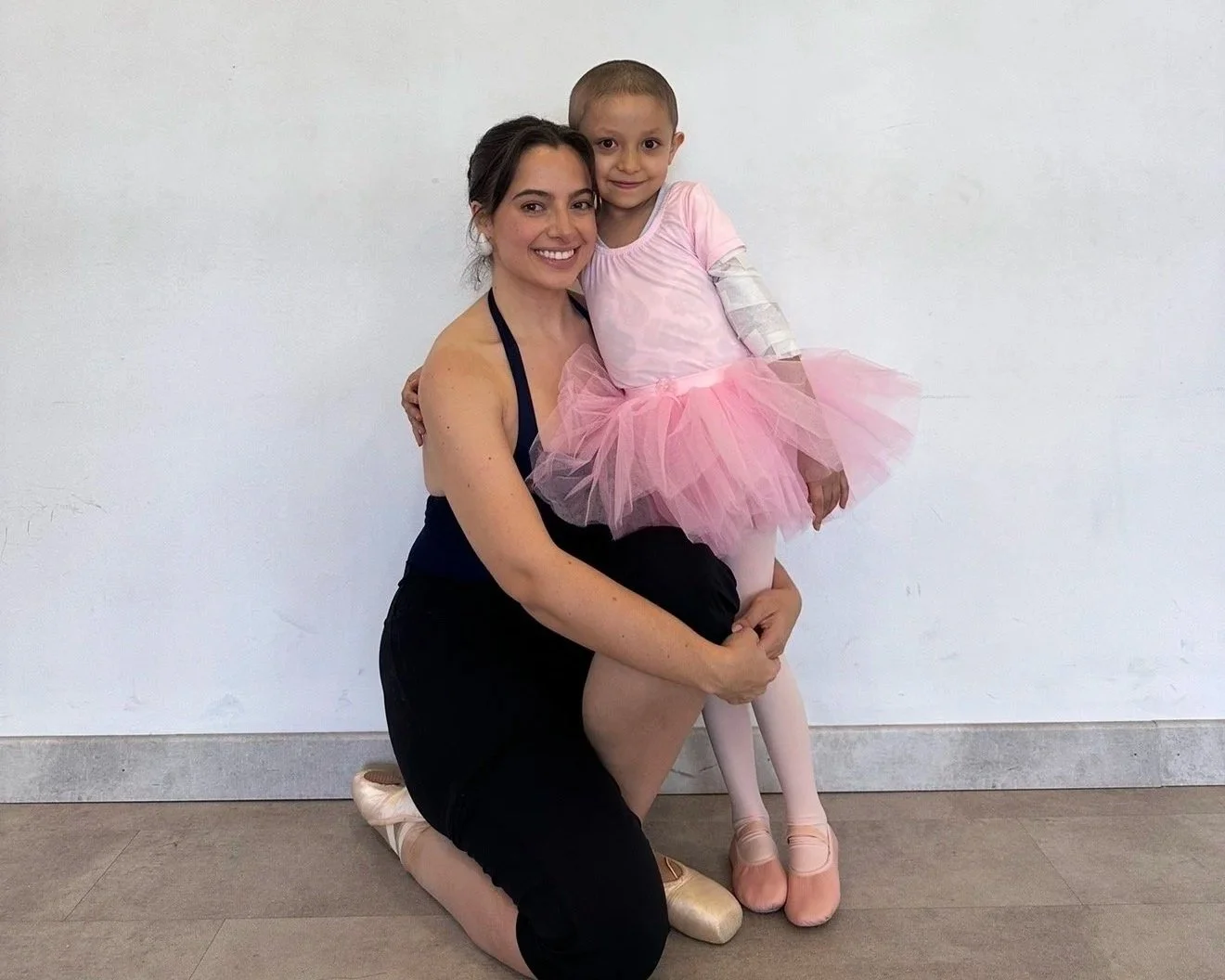 A ballerina in a black leotard and pink ballet slippers kneels next to a young girl in a pink tutu and ballet slippers, both smiling and posing against a plain white wall.