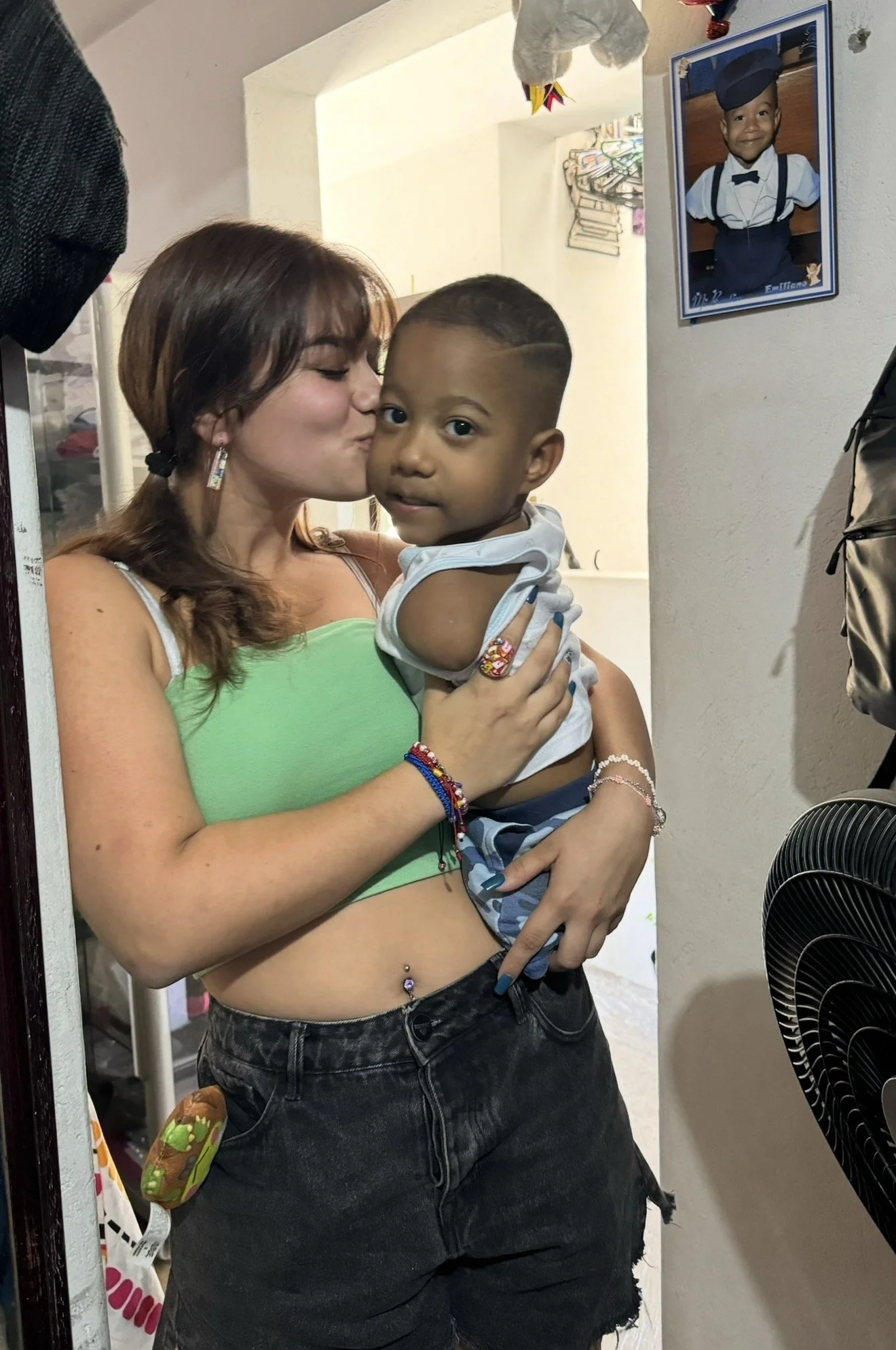 A woman is holding a young boy, giving him a kiss on the cheek inside a room. The woman has brown hair, is wearing a green top, and has various jewelry including earrings and bracelets. A framed photo of a child in a graduation cap and gown is hanging on the wall behind them.