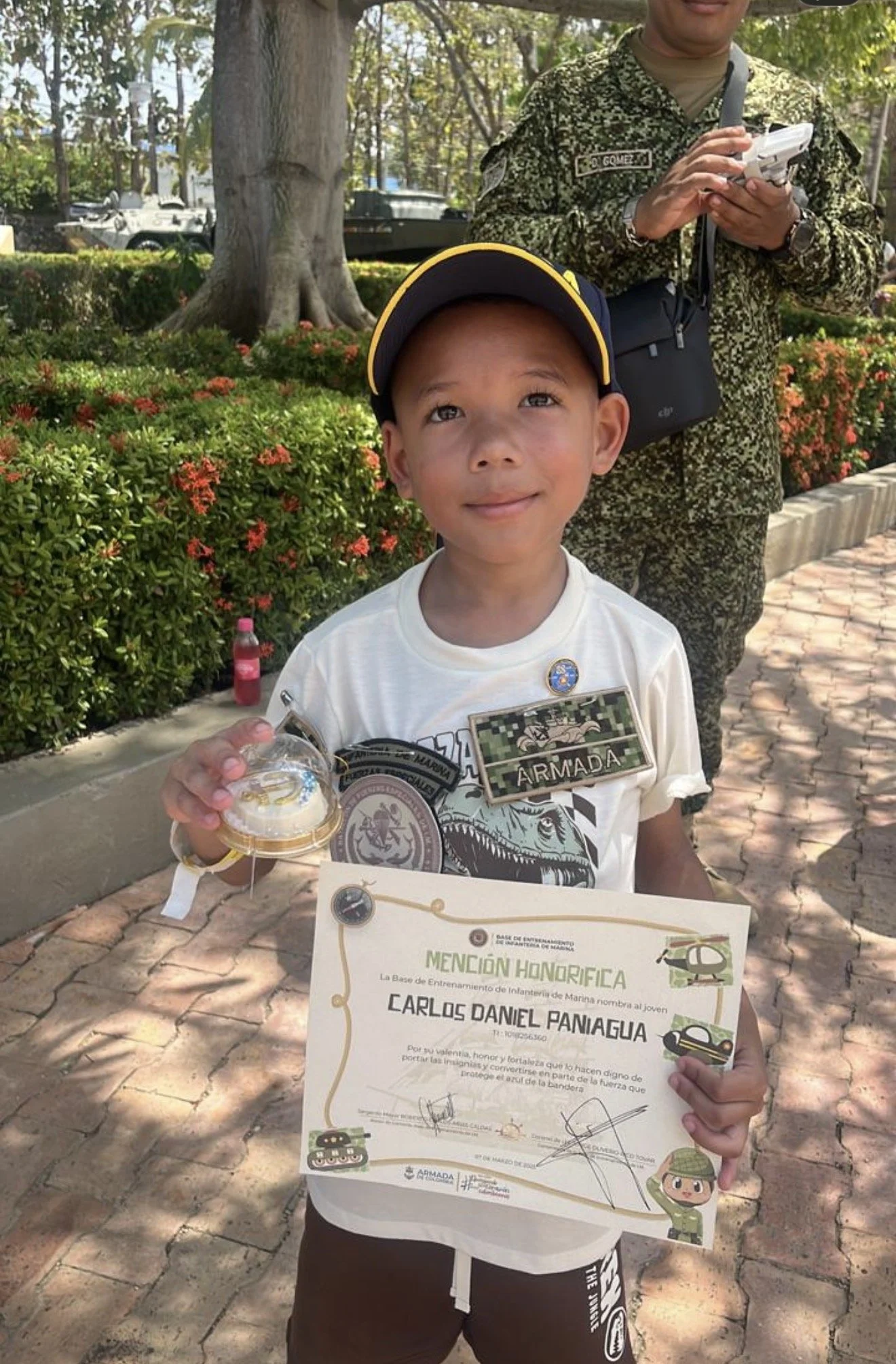 A young boy wearing a baseball cap and a white t-shirt holding a certificate of honor. An adult in military uniform stands behind him, holding a tablet. The boy is holding a small container, possibly a prize, and there is a water bottle on the ground nearby. The background features trees, flowers, and military vehicles.