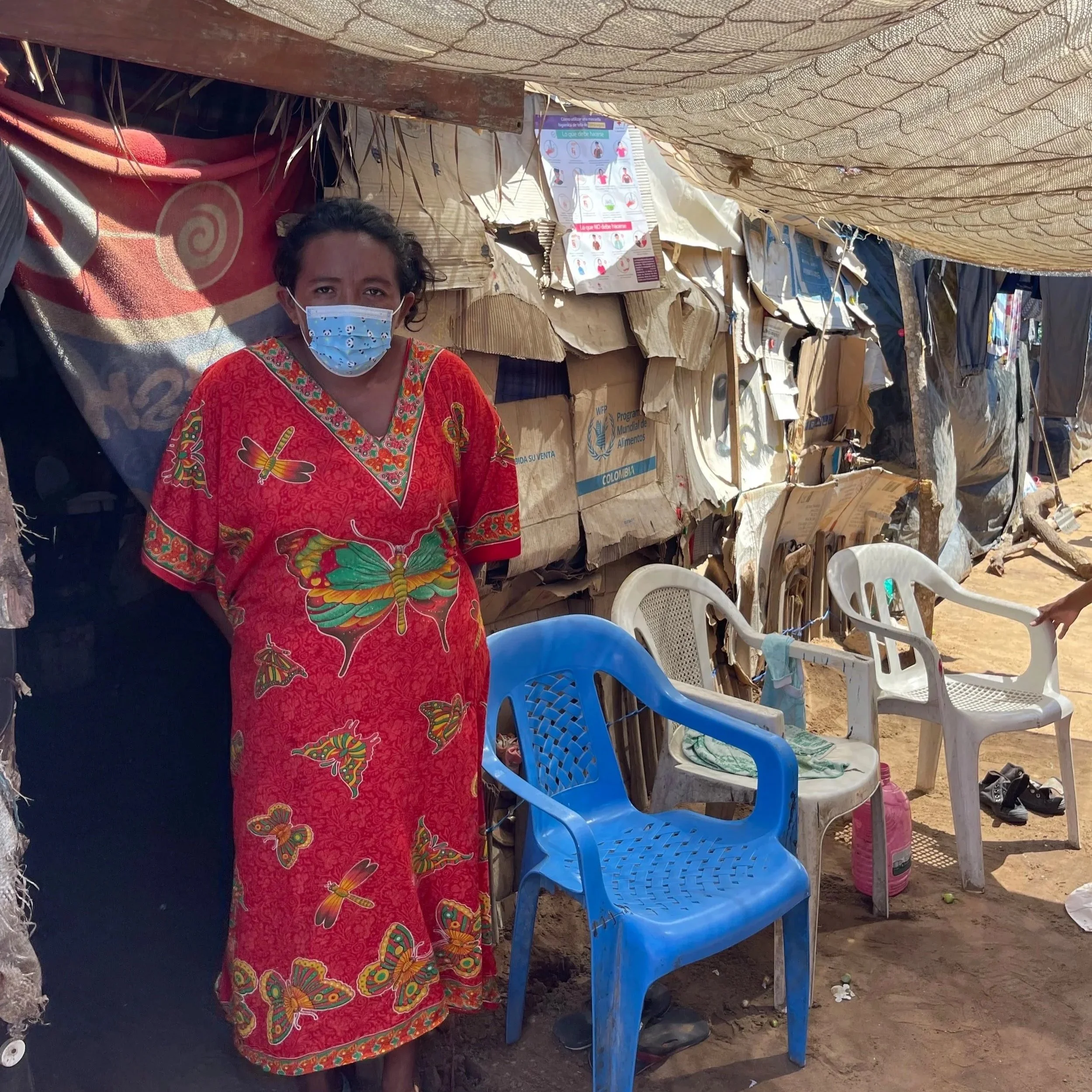A woman wearing a red dress with butterfly and floral patterns and a blue face mask standing next to plastic chairs and makeshift shelter made of cardboard and fabric in an outdoor setting.