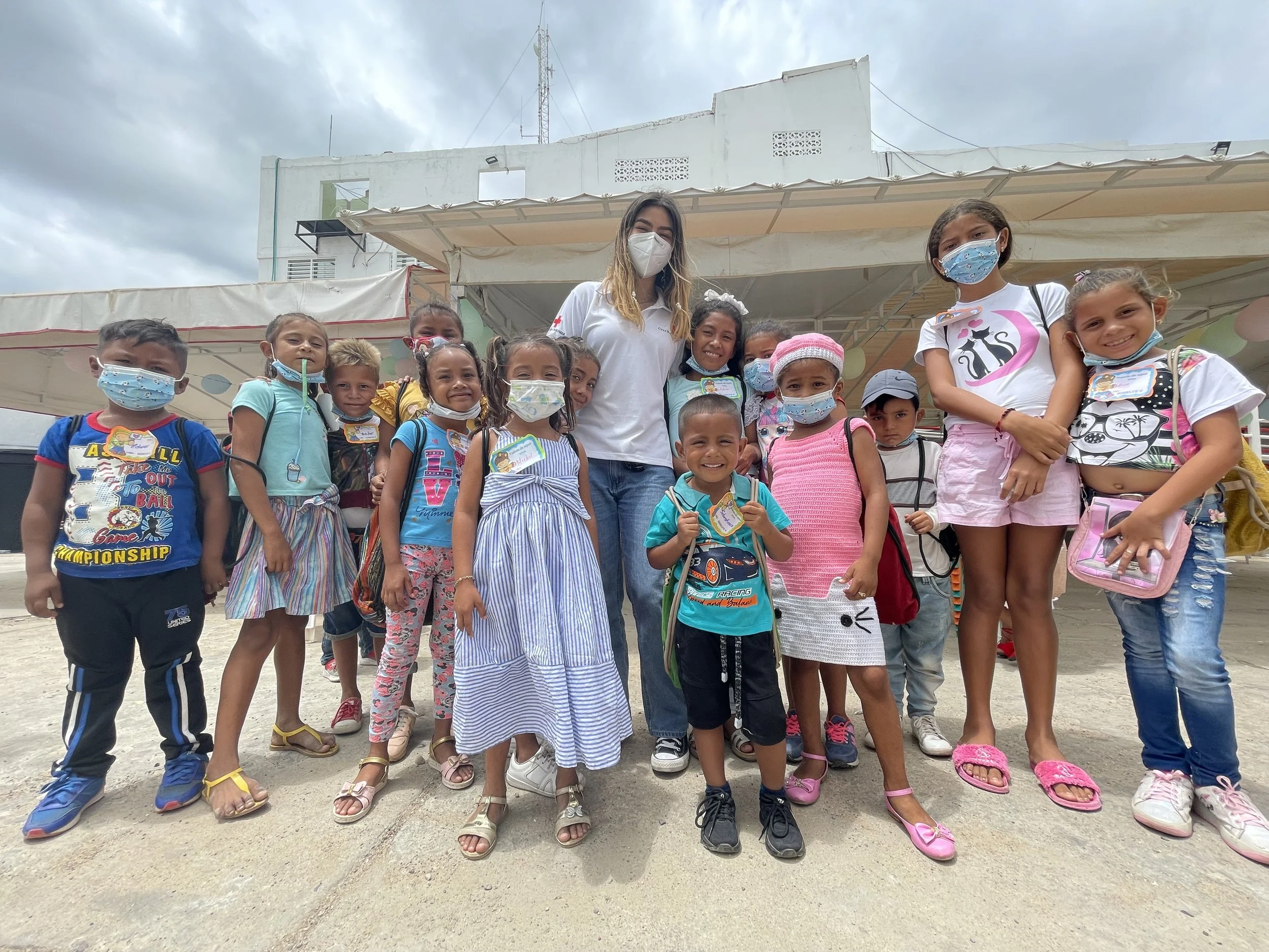 A group of children wearing face masks and colorful clothing, standing outdoors in front of a woman in a white shirt and face mask, with a cloudy sky overhead.