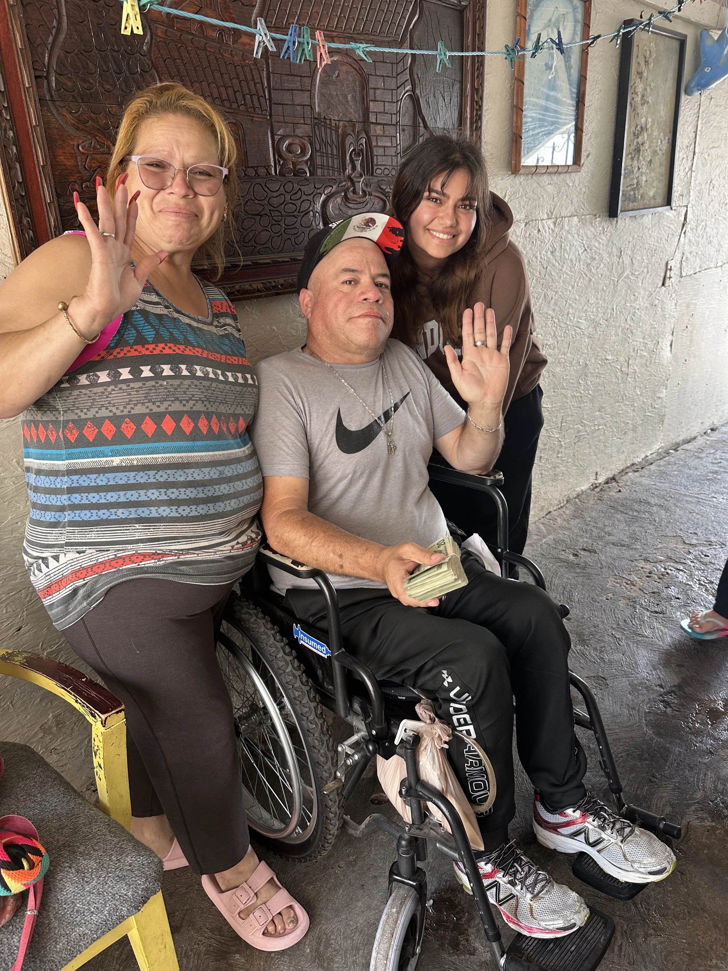 Three people posing for a photo: a woman with light skin, glasses, and long, red hair waving, a man with light skin in a wheelchair holding a stack of money, and a young woman with light skin and long dark hair, smiling and waving, standing behind them. They are indoors against a wall with artwork and hanging clothespins.