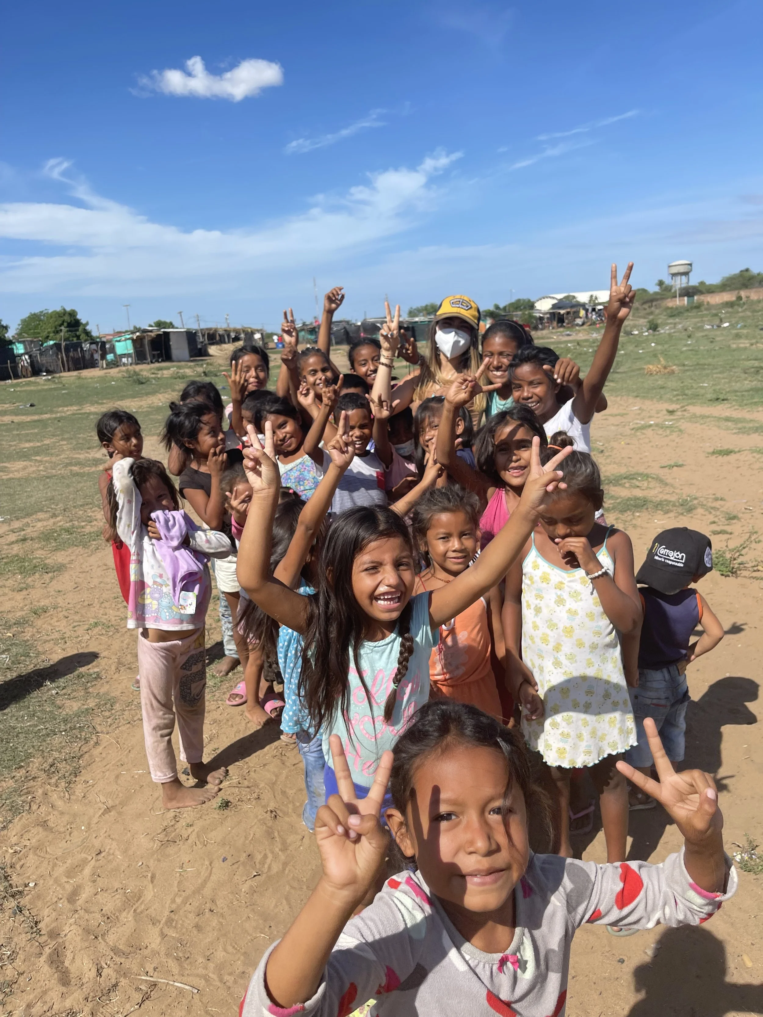 A group of smiling children standing outdoors on a sunny day, some making peace signs, with a dirt ground and structures in the background.