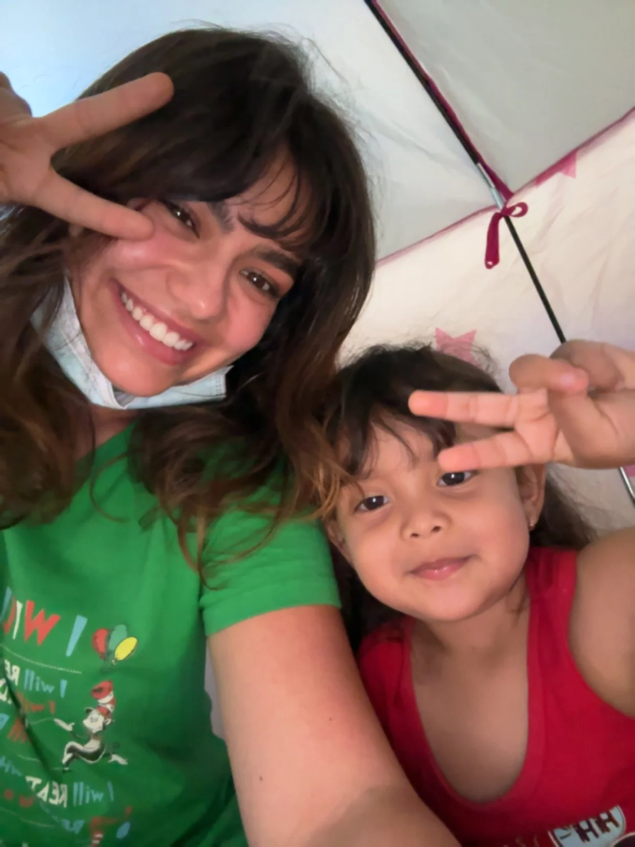 A woman and a young girl taking a selfie together, both making peace signs with their hands. The woman is smiling widely and has brown hair, while the girl is also smiling and has dark hair, wearing a red shirt. They are inside a tent with a pink ribbon and star pattern.