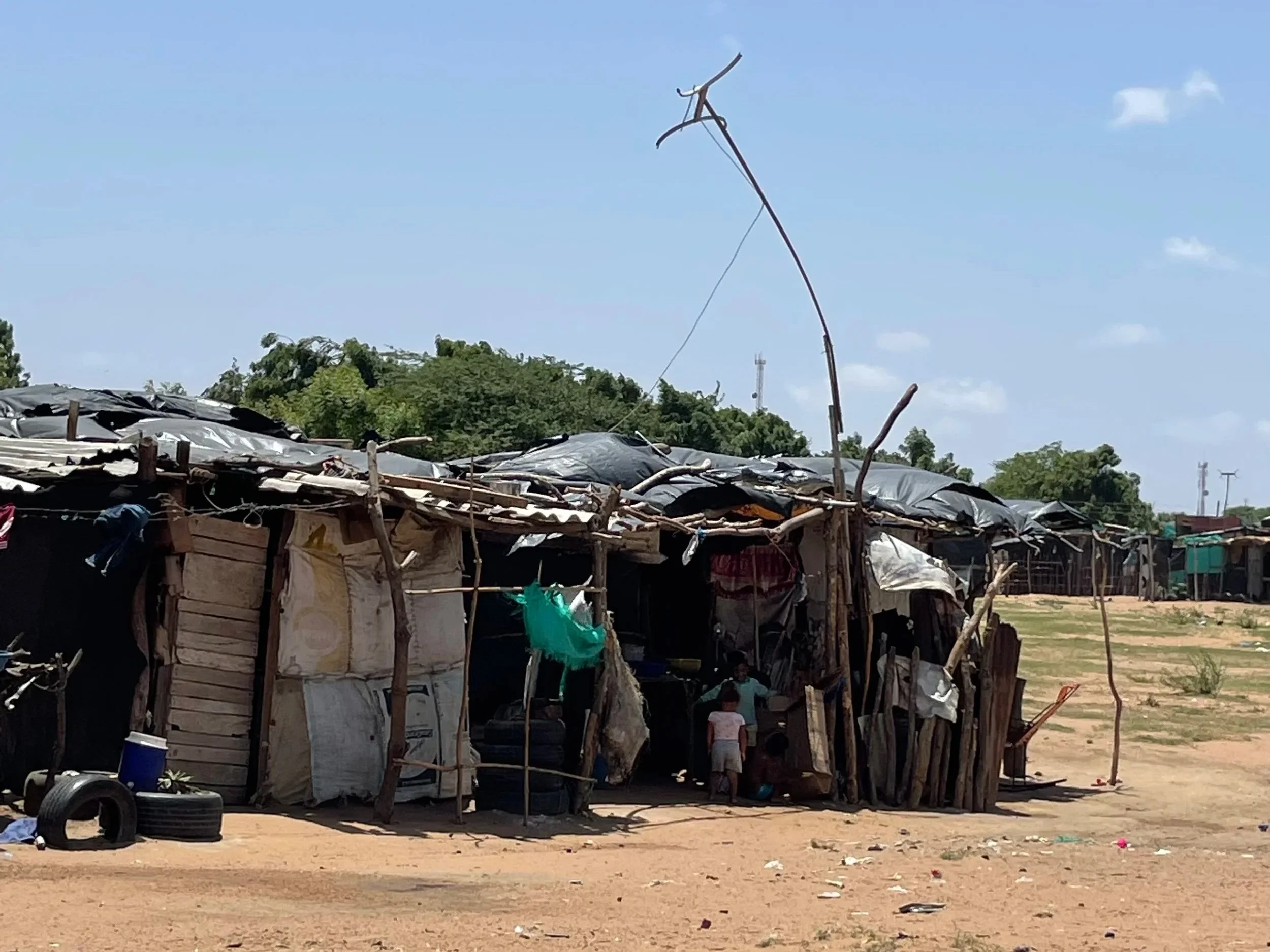A makeshift shelter made of wood and plastic sheets, with children standing nearby, in a rural area with trees and a partly cloudy sky.