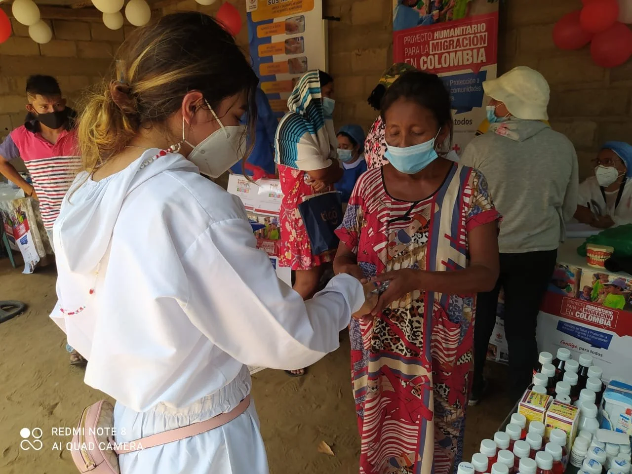 A woman in a white jacket and medical mask hands a small item to an older woman wearing a colorful patterned dress and medical mask at a health outreach event with tables of supplies, posters, and other people in masks in the background.