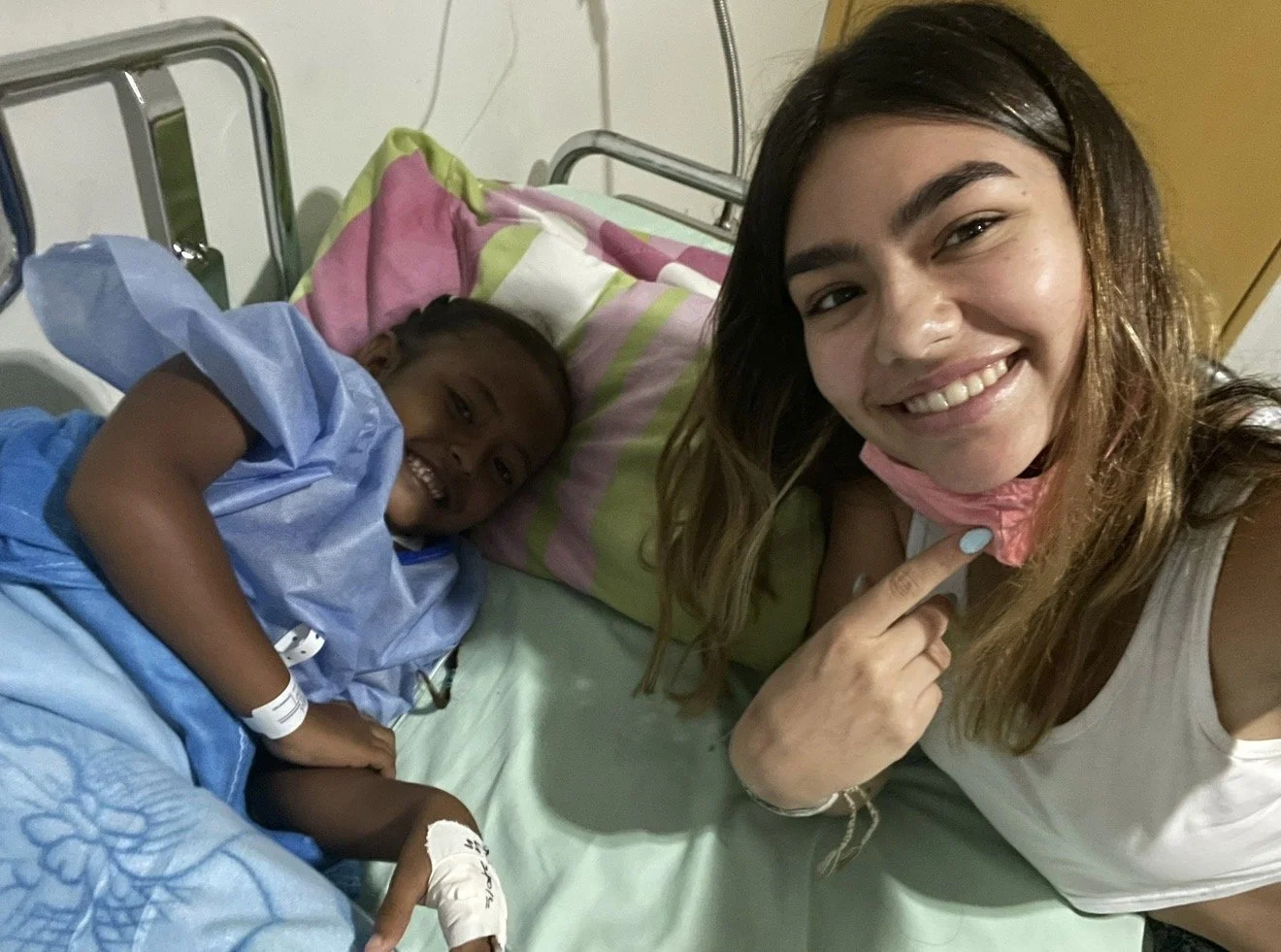 A woman smiling and pointing to her face taking a selfie with a young girl lying in a hospital bed, smiling and holding her hand, both in hospital attire.
