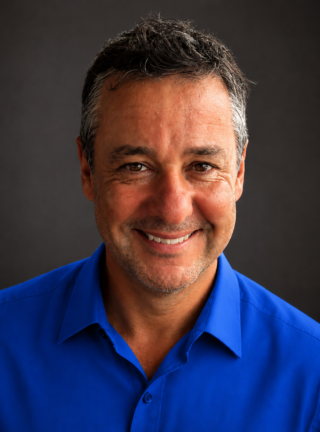 A middle-aged man with short, dark hair, some gray, smiling, wearing a bright blue button-up shirt, against a dark background.
