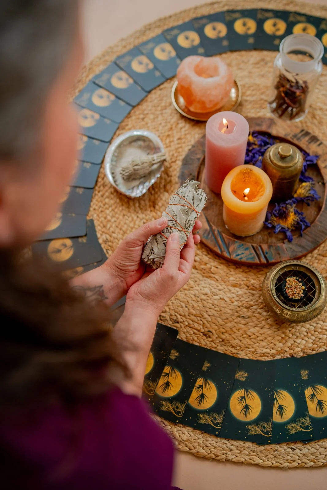 Eine Person hält eine Bündel aus getrockneten Kräutern über einem Tisch mit Kerzen, Kristallen, und Tarotkarten. Der Tisch ist mit einem Rattan-Tuch bedeckt, umgeben von verschiedenen Ritual- und Zauberutensilien.