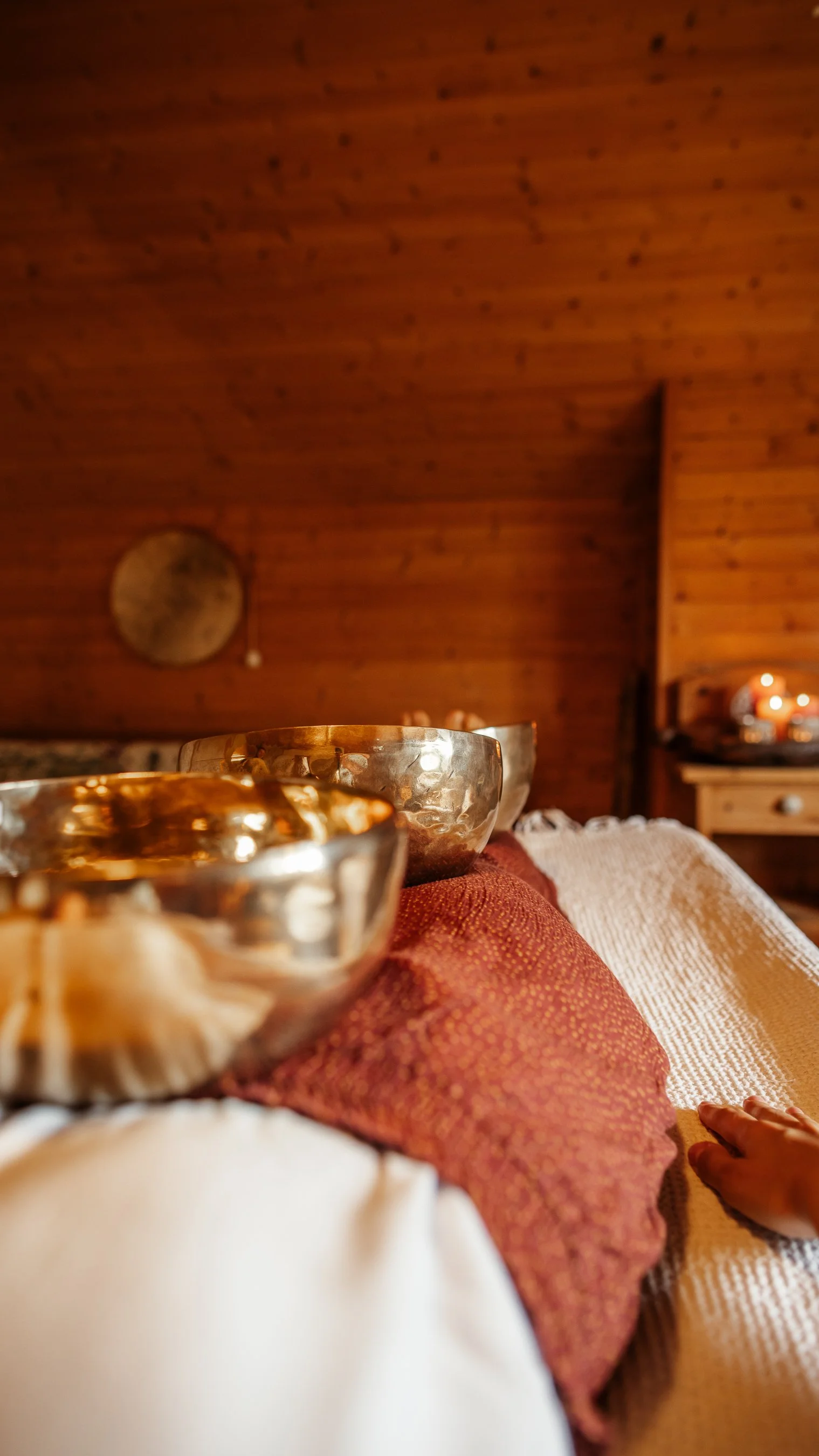 Silberne Klangschalen liegen auf einer roten und weißen Decke in einem gemütlichen Raum mit Holzvertäfelung und Kerzenlicht.