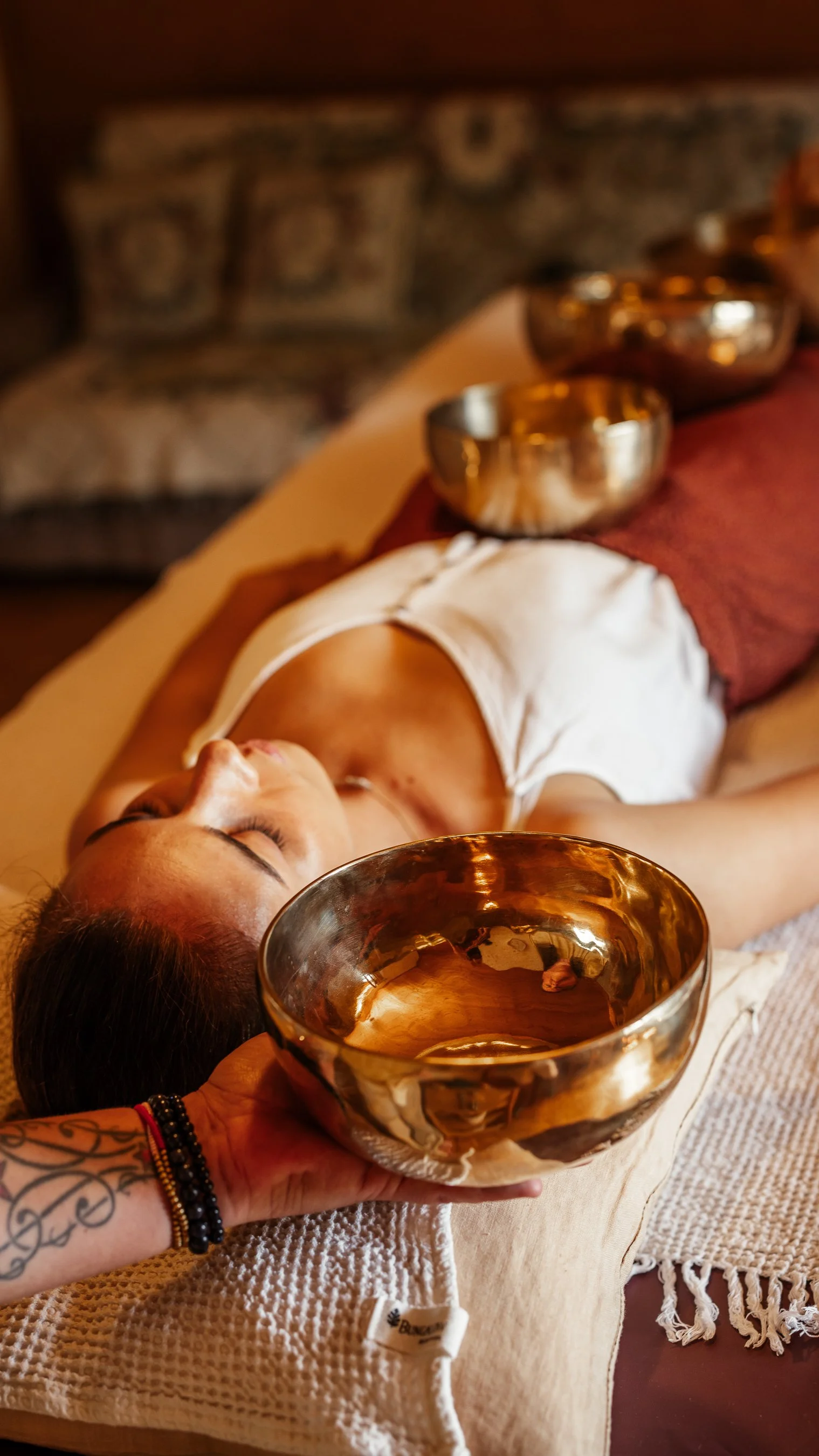 Eine Frau liegt entspannt auf einer Massagebank, während eine Hand mit Tattoo und Armbändern mit einer goldfarbenen Schüssel im Vordergrund eine Wasserzeremonie durchführt.