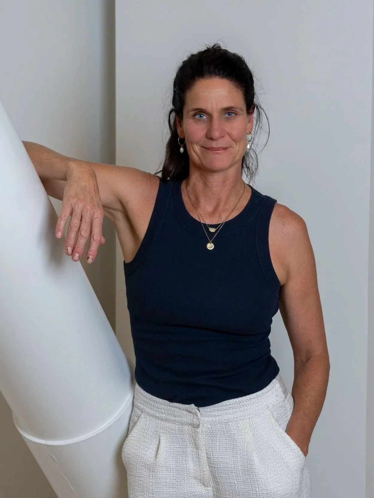 A woman with dark hair, blue eyes, wearing a black sleeveless top and white textured pants, stands indoors next to a white column, smiling softly.
