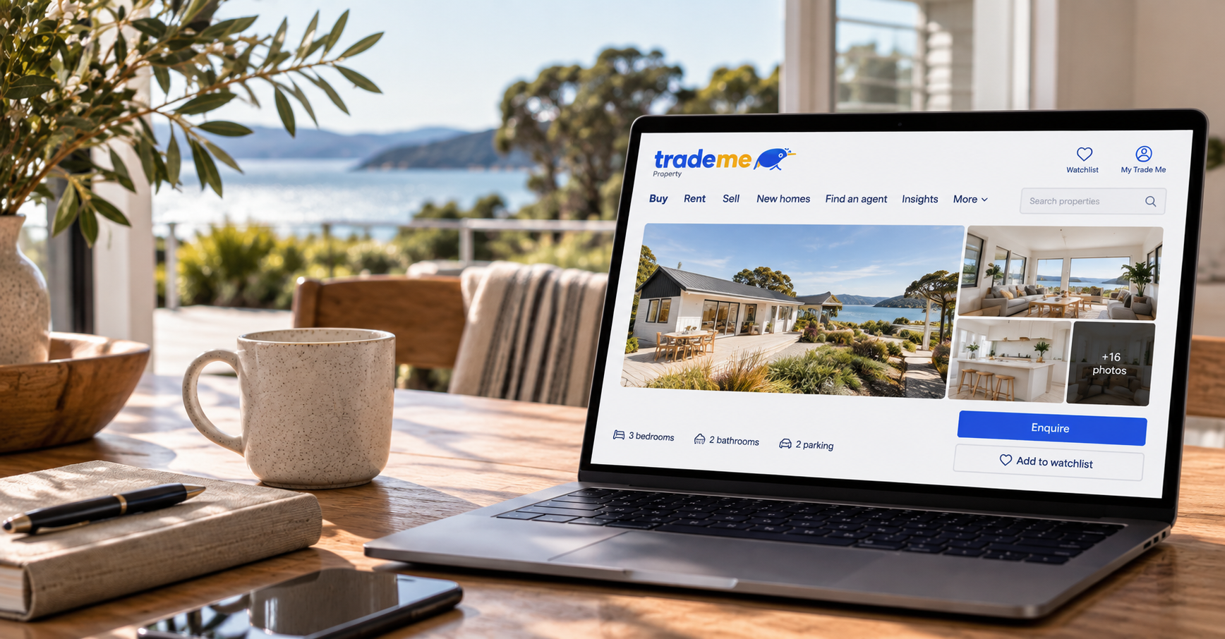 A laptop displaying a real estate listing for a house with a view of water, located on a wooden table inside with a mug, pen, notebook, and smartphone nearby, overlooking a deck and outdoor scenery.